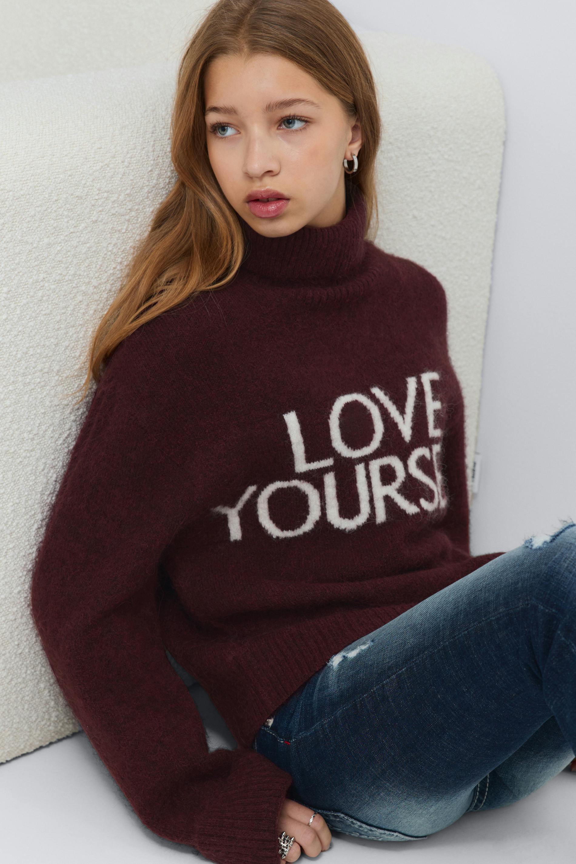 Burgundy fuzzy turtleneck sweater with white "LOVE YOURSELF" lettering, paired with distressed blue jeans and silver hoop earrings and rings.