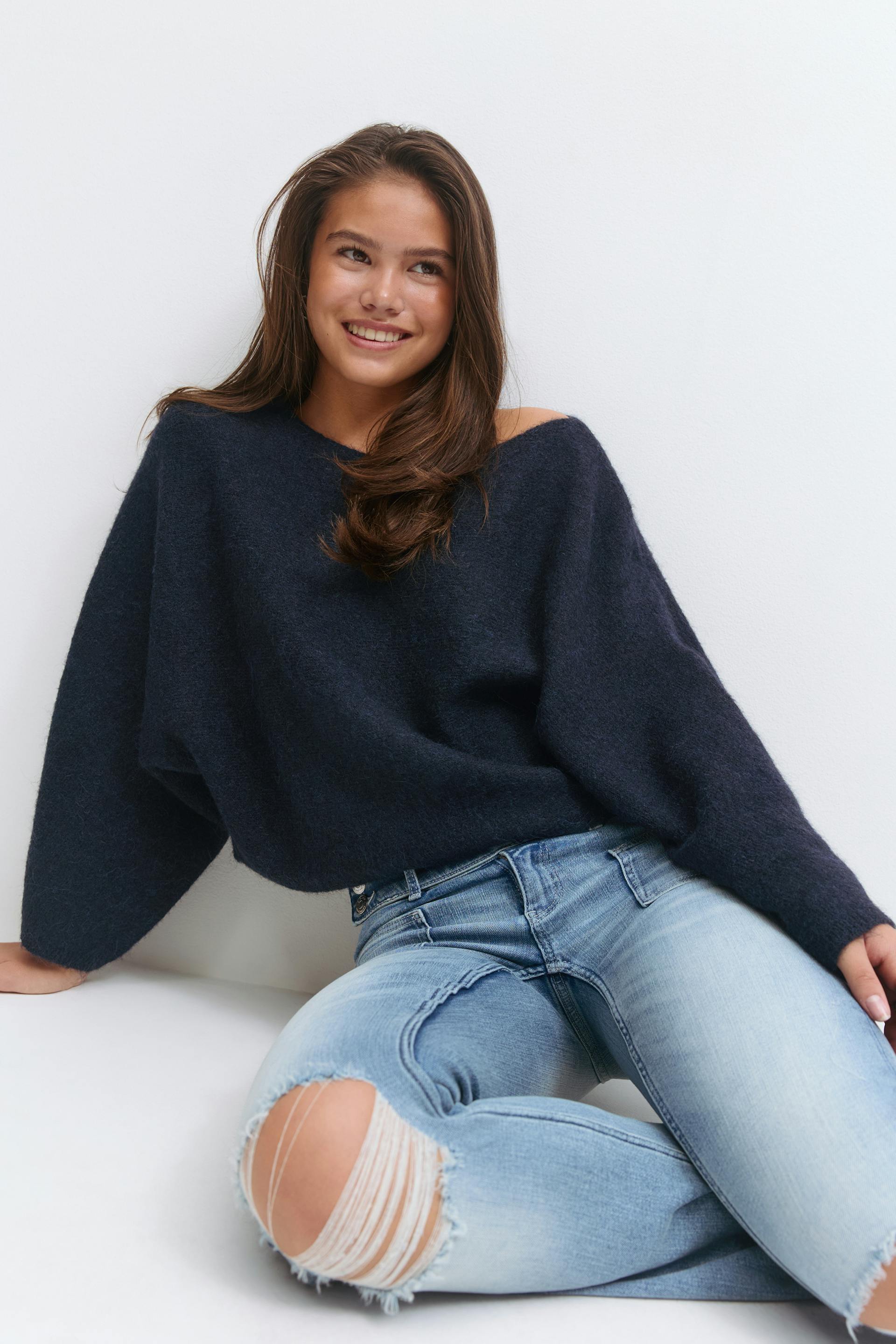 Navy off‑shoulder oversized fuzzy sweater paired with light‑wash distressed skinny jeans featuring a large frayed knee rip.