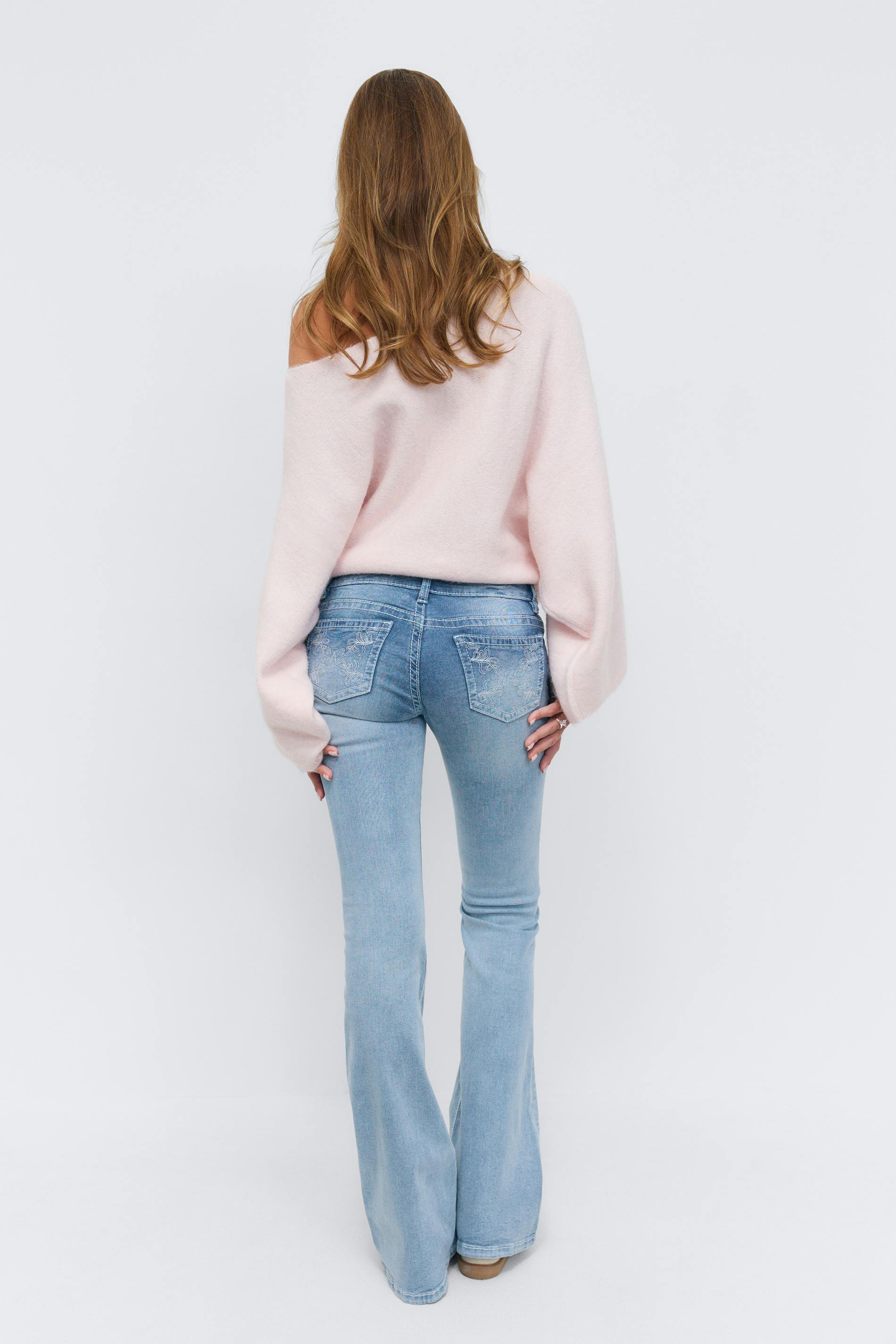 Light pink off-the-shoulder sweater with long, voluminous sleeves, paired with light‑wash flared jeans featuring embroidered back pockets.