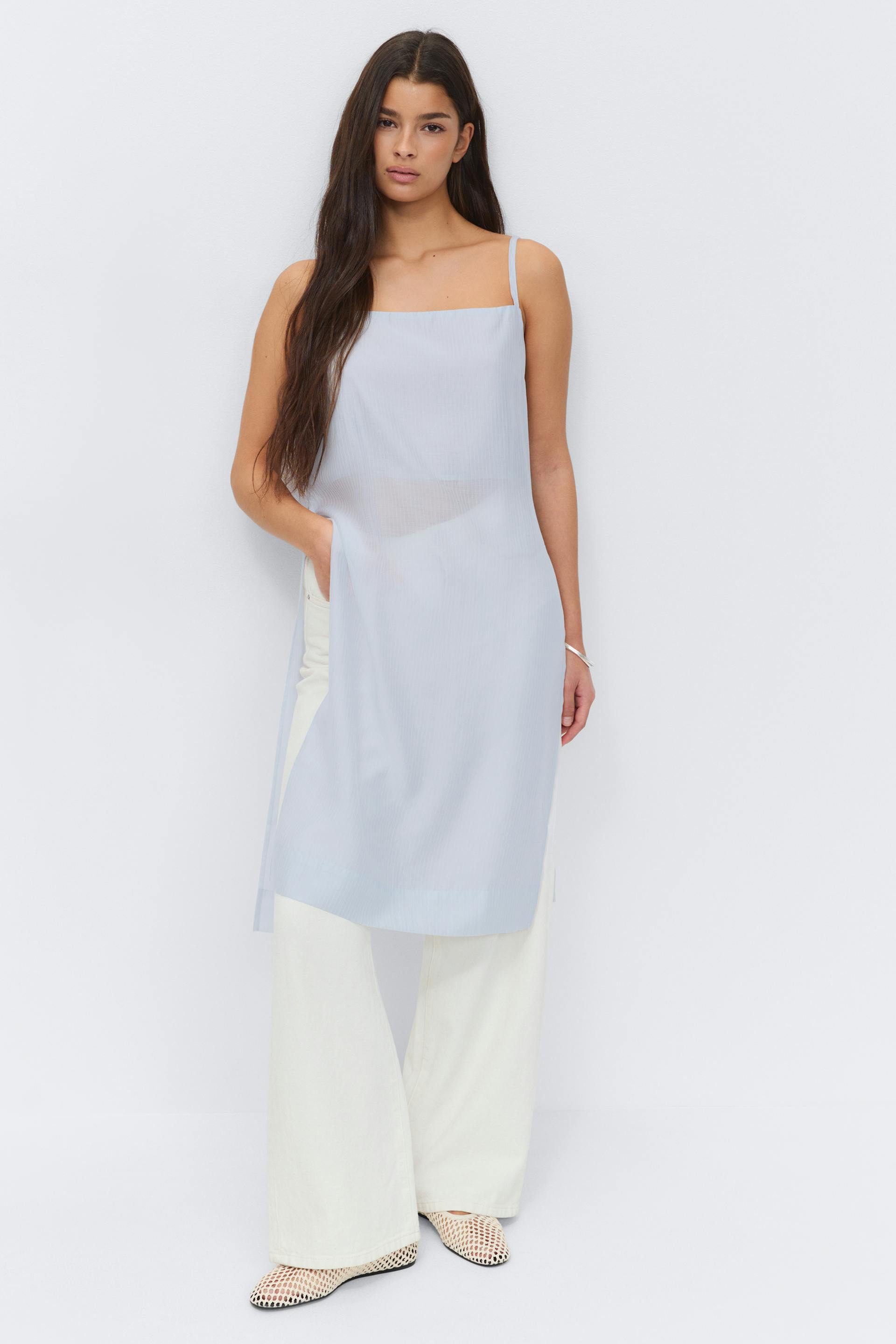 Hellblaue ärmellose Tunika mit dünnen Schulterträgern über einer weißen Hose mit weitem Bein, kombiniert mit weißen gewebten, spitzen Schuhen.