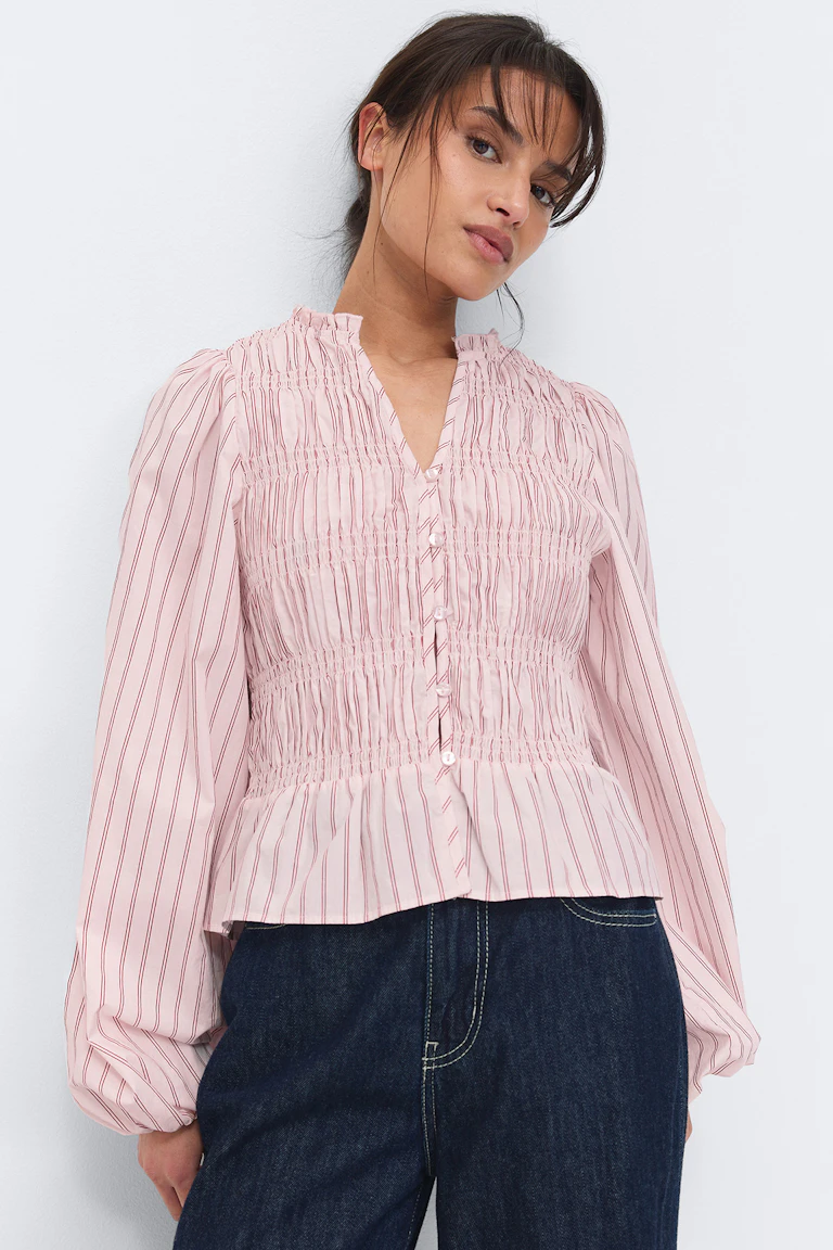 Ljusrosa blus med röda vertikala ränder, smockat ok och volangkrage, V-ringad hals med en knapp och voluminösa ballongärmar som är rynkade i elastiska muddar, kombinerad med mörkblå jeans.