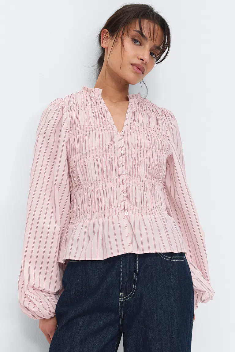 Lys rosa bluse med røde vertikale striper, smoket bærestykke og volangkrage, V-utringning med én knapp og voluminøse ballongermer med elastiske mansjetter, sammen med mørkeblå jeans.