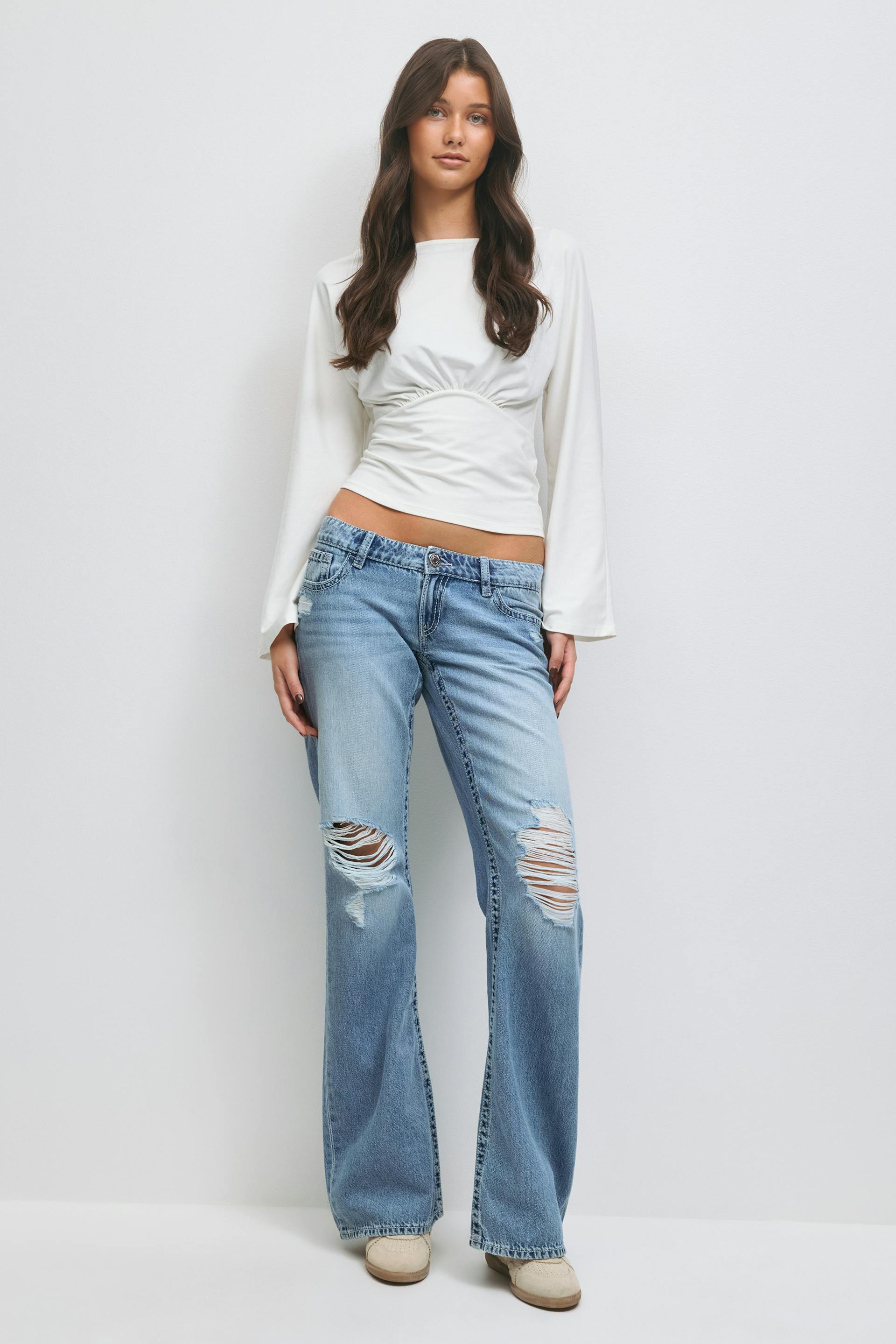 Bijgesneden witte getailleerde top met lange mouwen, rimpel onder de buste en iets uitlopende manchetten; licht verwassen, uitlopende jeans met grote scheuren in de knie; beige slip-on schoenen.