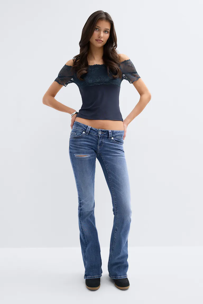 Gekürztes, cremefarbenes, geripptes, langärmeliges Oberteil mit Schlitz auf der Vorderseite, gepaart mit einer mittelhohen, dunkel gewaschenen Skinny-Jeans mit horizontalem Riss am Oberschenkel; einfache Silberringe an der linken Hand.