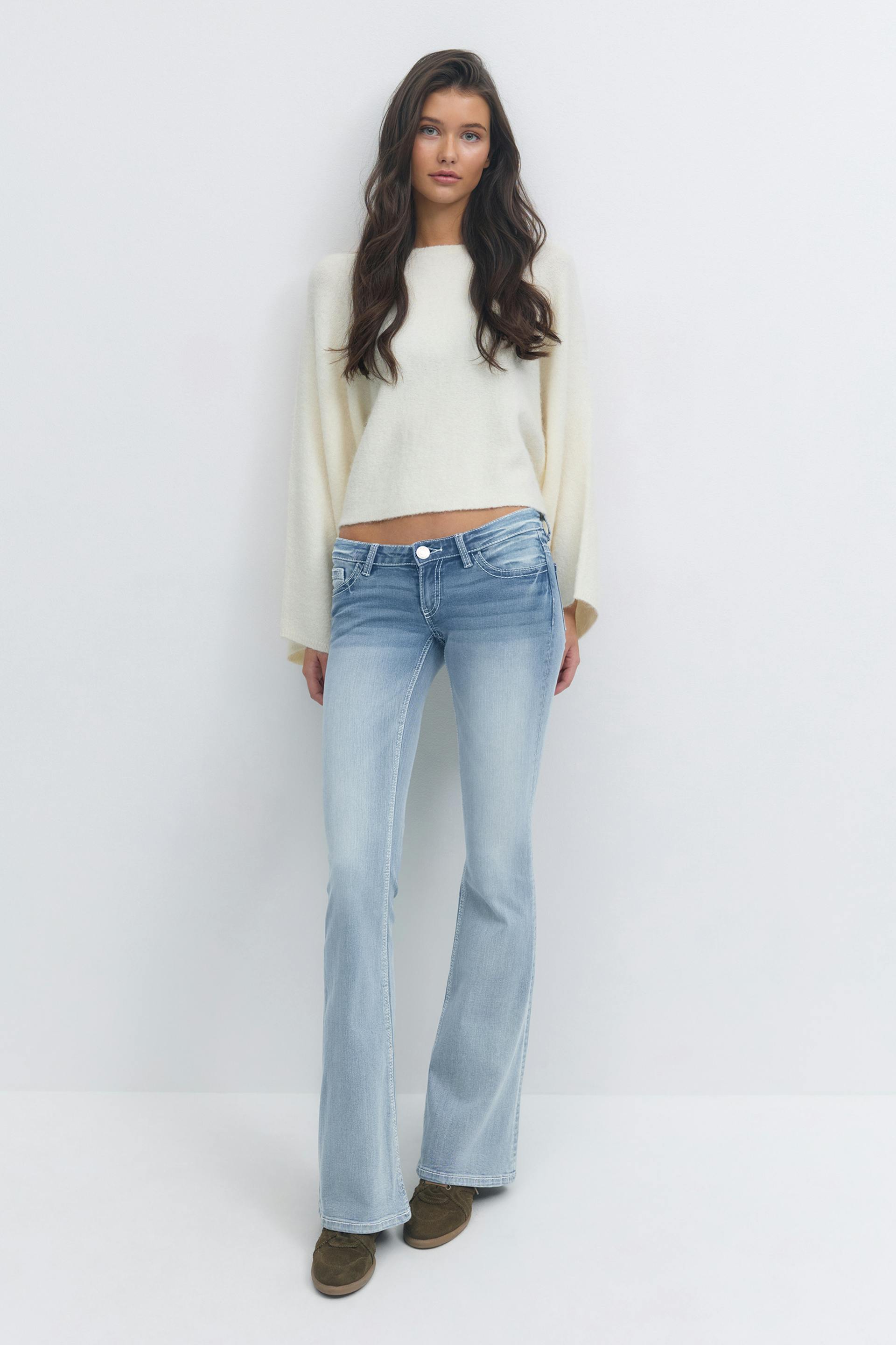 Ivoorkleurige korte trui met lange mouwen, lichtgekleurde bootcut jeans en bruine suède veterschoenen.