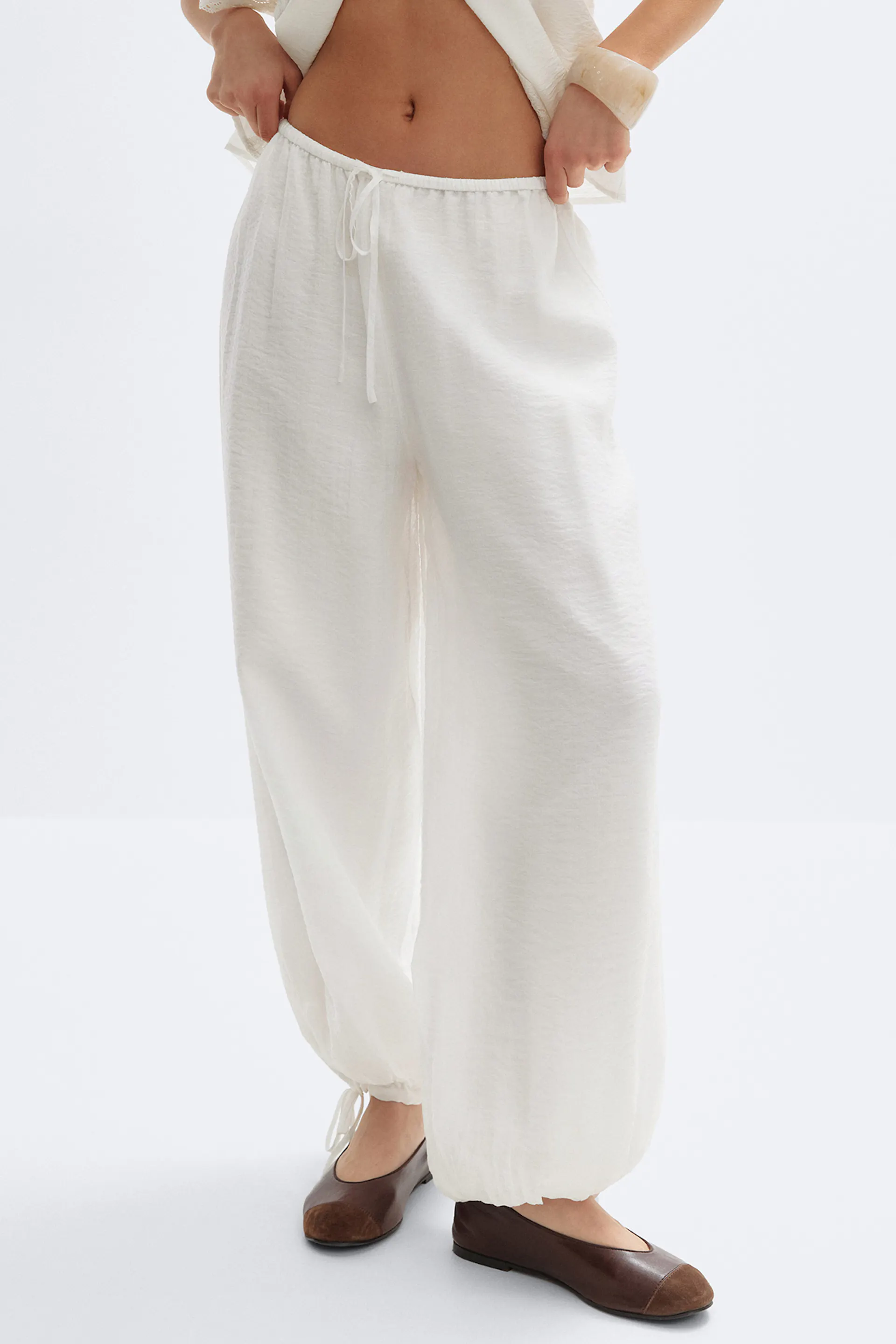 Cremeweiße Hose mit weitem Bein und Gummizug an den Knöcheln, kombiniert mit braunen Lederballets mit dunkleren Zehenkappen.