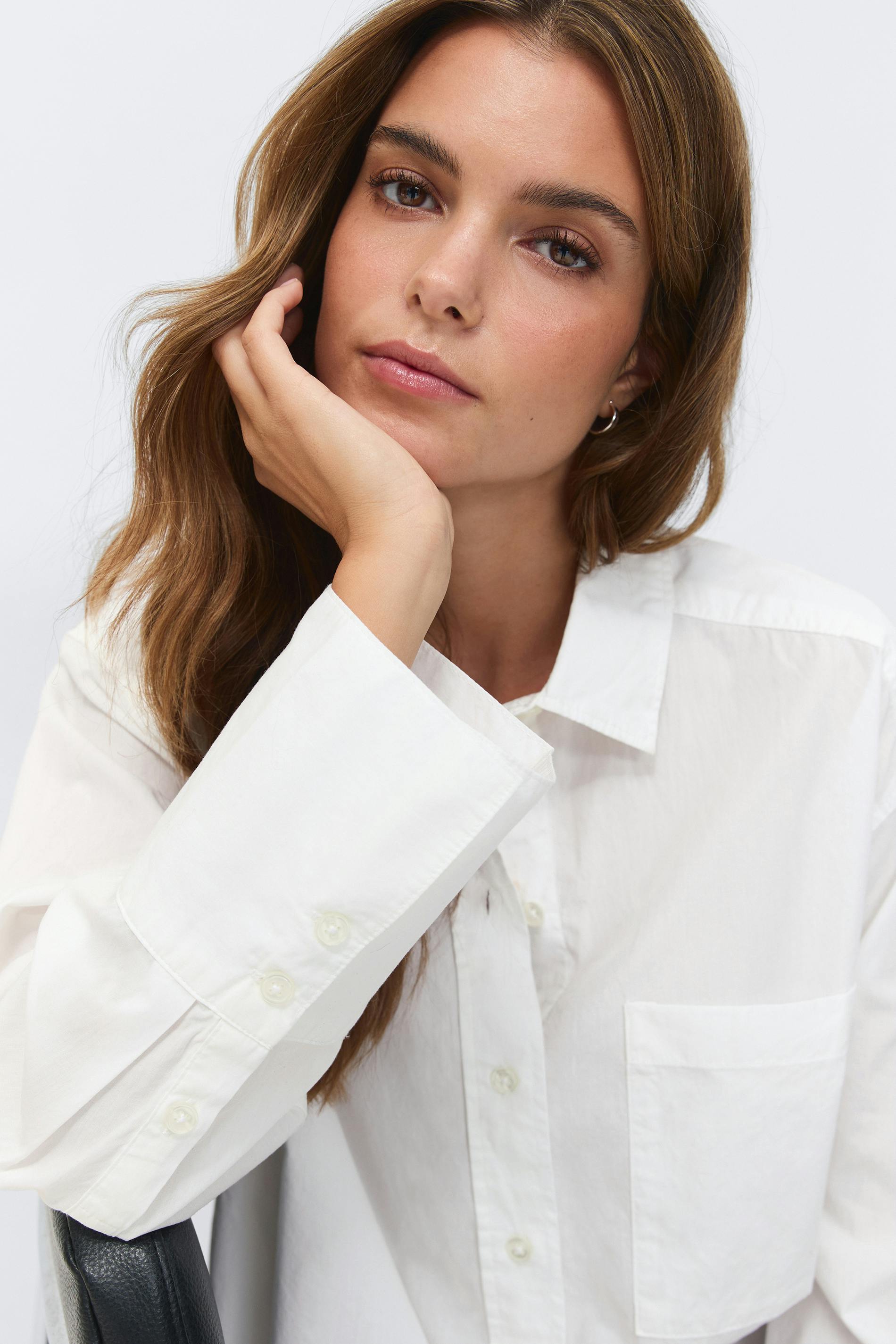 White oversized long-sleeve button-down shirt with chest pocket, styled with light-wash high-rise straight-leg jeans and small gold hoop earrings.