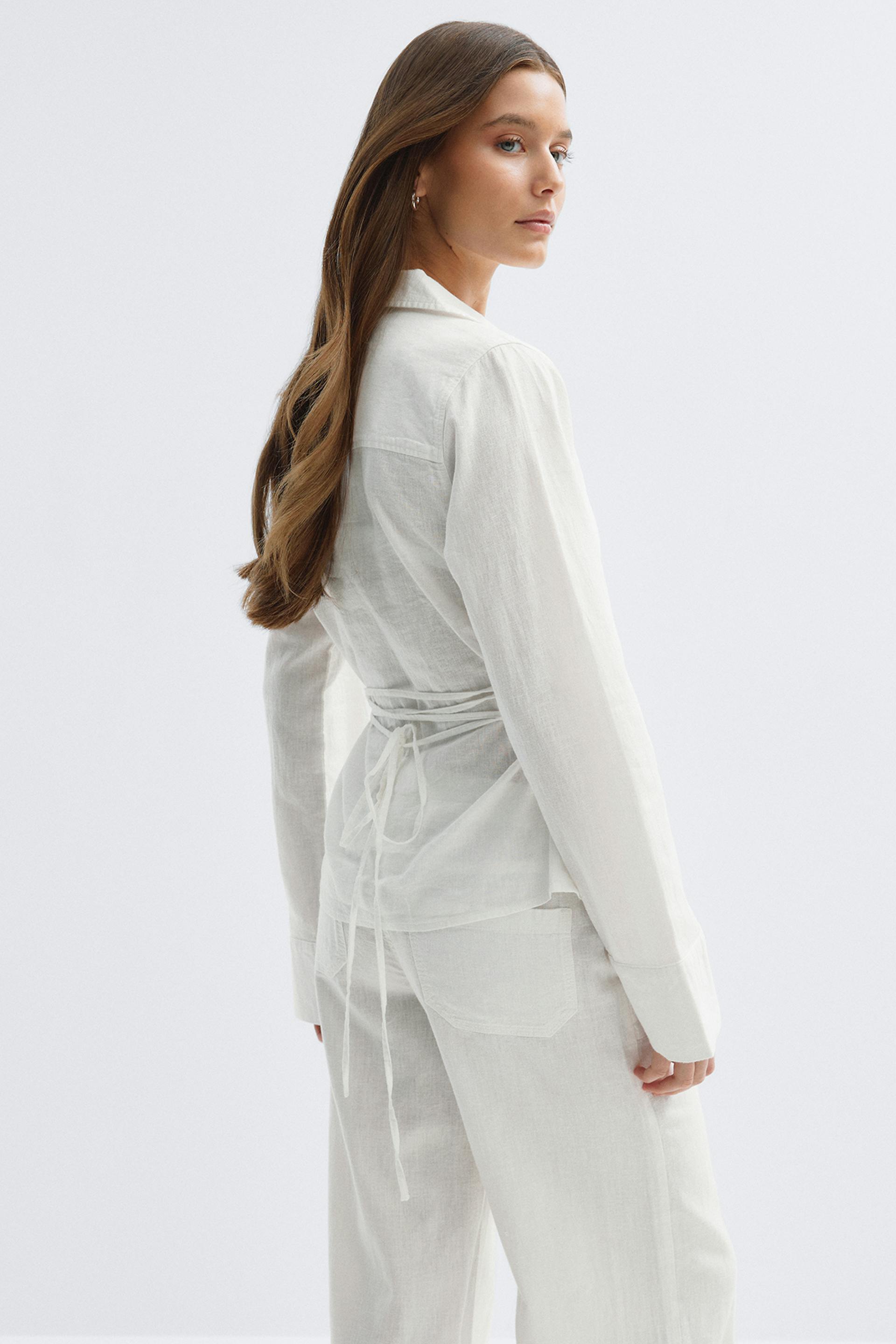 Chemisier à manches longues en lin léger blanc avec détails à nouer, associé à un pantalon en lin blanc à la coupe décontractée avec poches plaquées à l'arrière.