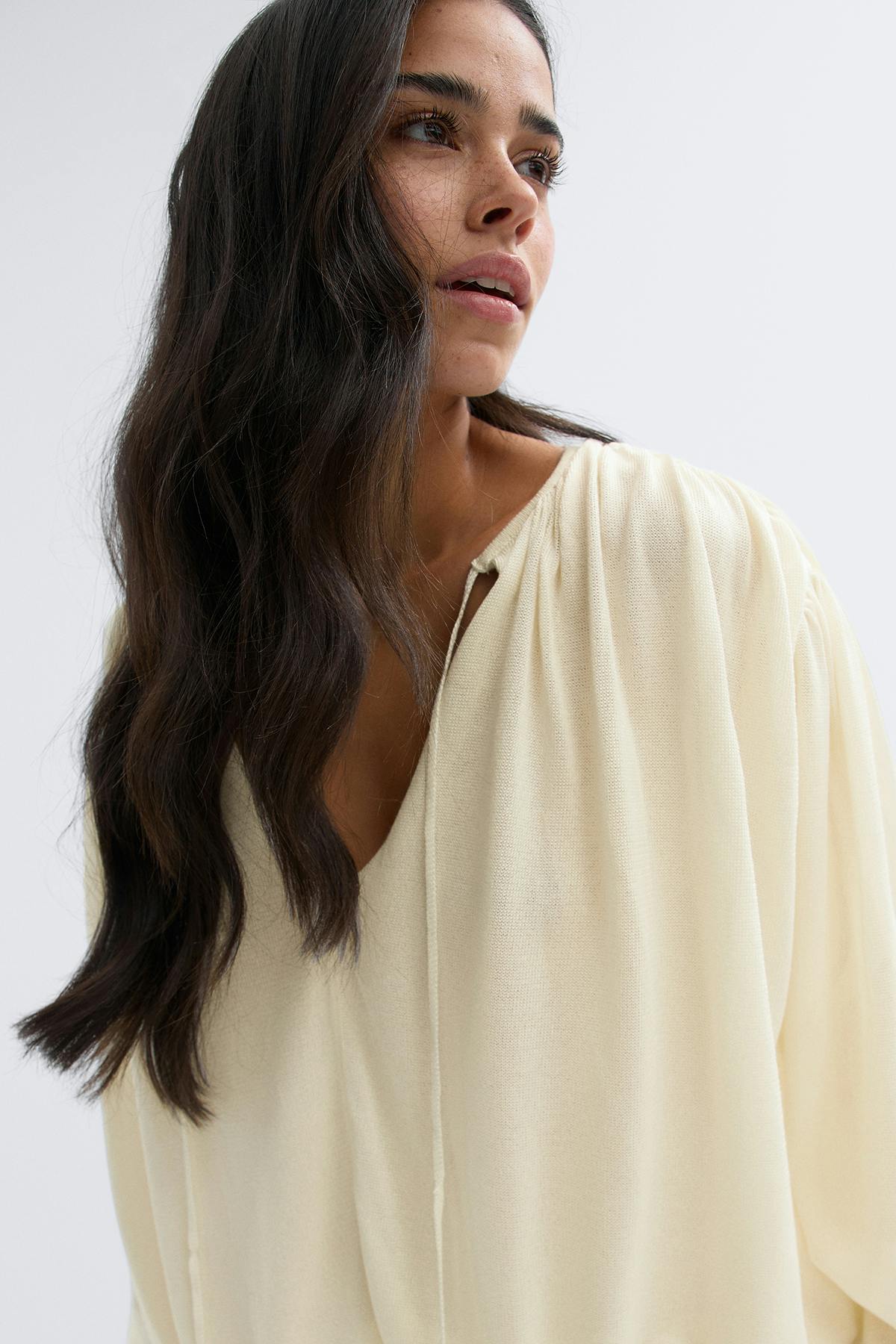 Kermanvärinen pitkähihainen V-kauluksinen pusero, jossa solmiopääntie ja tuuheat hihat, löysästi pujotettu korkeavyötäröisiin vaaleisiin leveälahkeisiin farkkuihin; mustat teräväkärkiset lenkkarit.