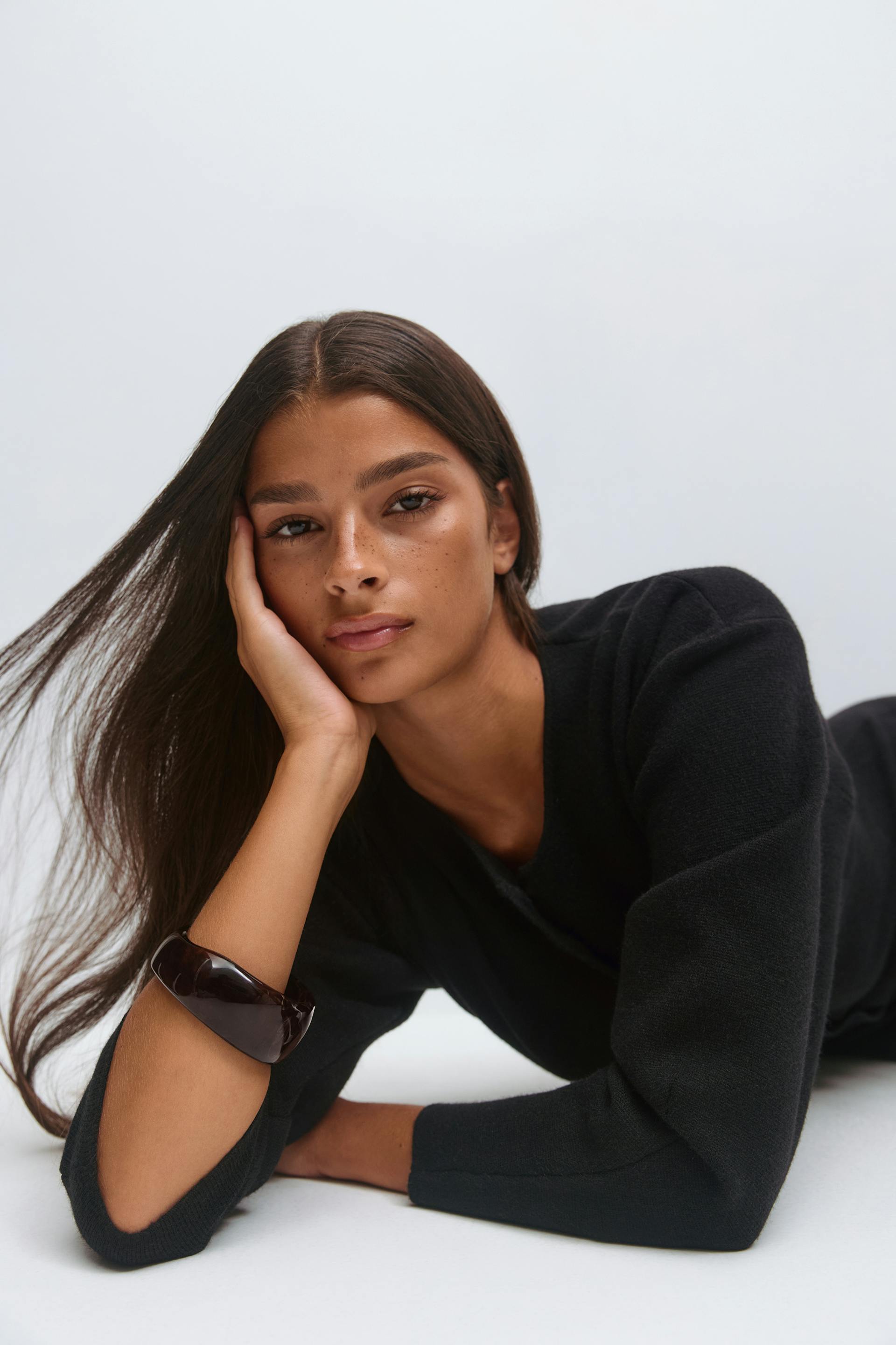 Musta pitkähihainen neuletoppi, jossa on pyöreä kaula-aukko ja hienovaraisesti paisutetut olkapäät, yhdistettynä suureen tumman kiiltävään kilpikonnarannekoruun.