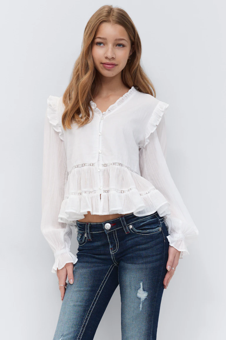 Weiße, langärmelige Schößchenbluse mit Knopfleiste, Rüschenbesatz, Spitzeneinsätzen und gerafften Manschetten. Dazu passt eine Skinny Jeans in dunkler Waschung mit Kontrastnähten und einem Used-Patch am Oberschenkel.