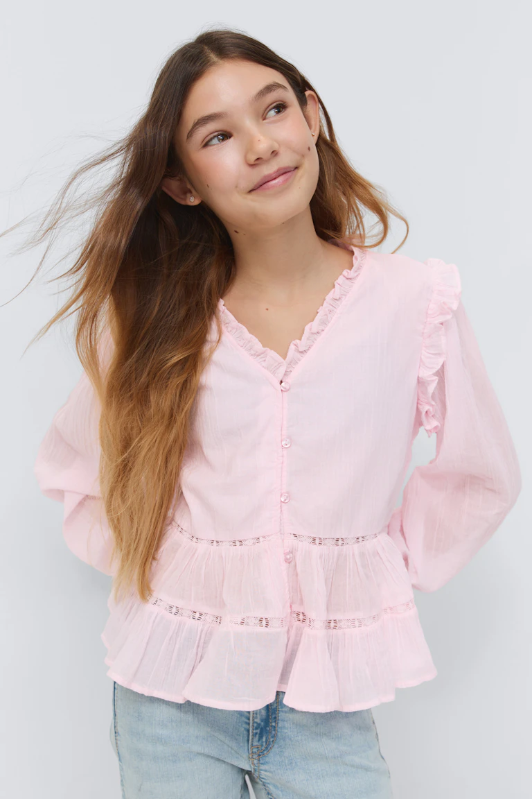 Chemisier rose pâle à manches longues boutonnées sur le devant, avec encolure en V volantée, manches bouffantes et péplum en dentelle, associé à un jean délavé.