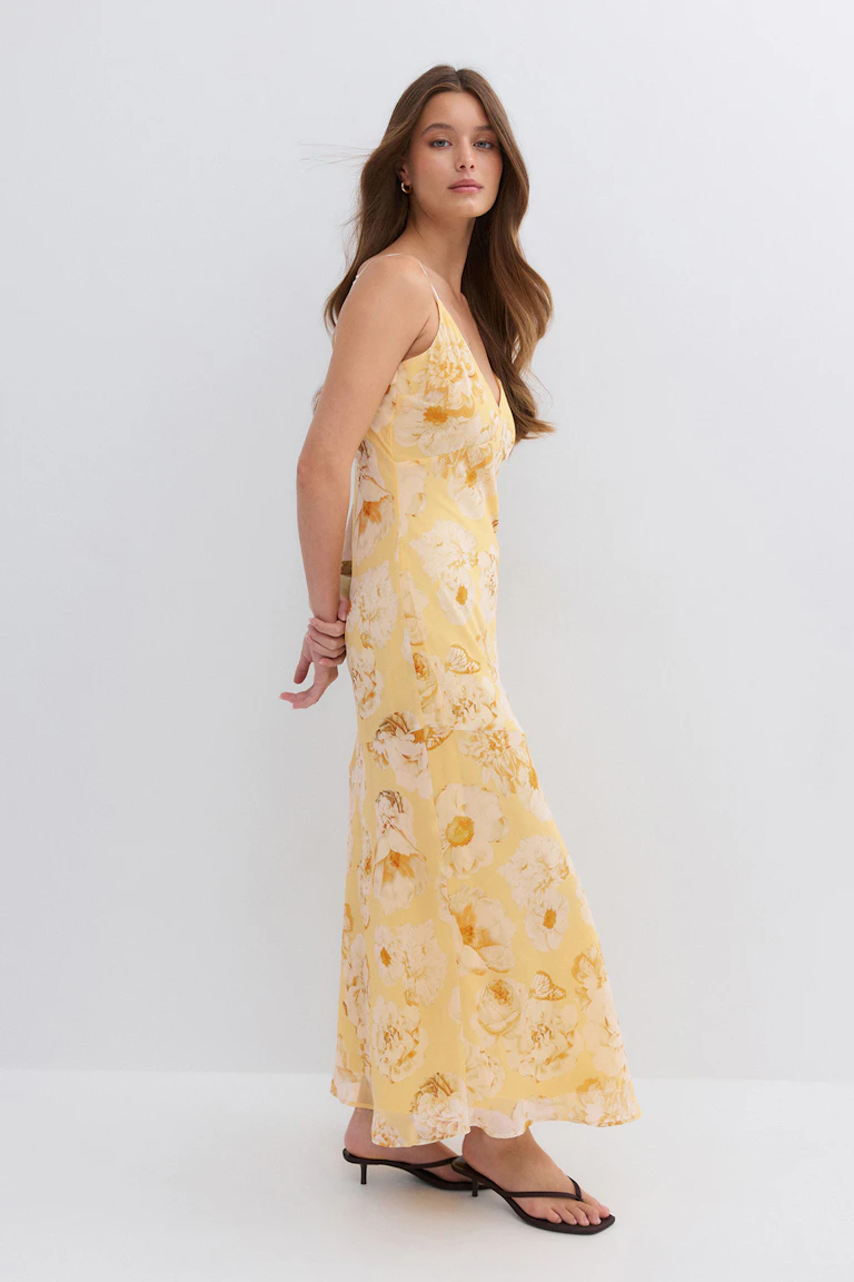 Gelbes, geblümtes Maxikleid mit V-Ausschnitt und dünnen Spaghetti-Trägern, kombiniert mit schwarzen Kitten-Heel-Sandalen und kleinen goldenen Ohrringen.