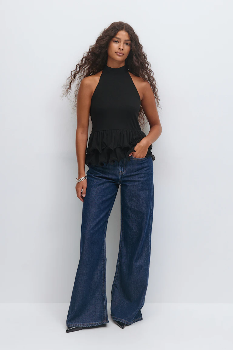 Schwarzes ärmelloses Neckholder-Top mit hohem Ausschnitt und mehrlagigem Rüschenschößchen; dunkle indigoblaue Jeans mit weitem Bein; schwarze, spitze Absätze und silberne, gestapelte Armbänder.