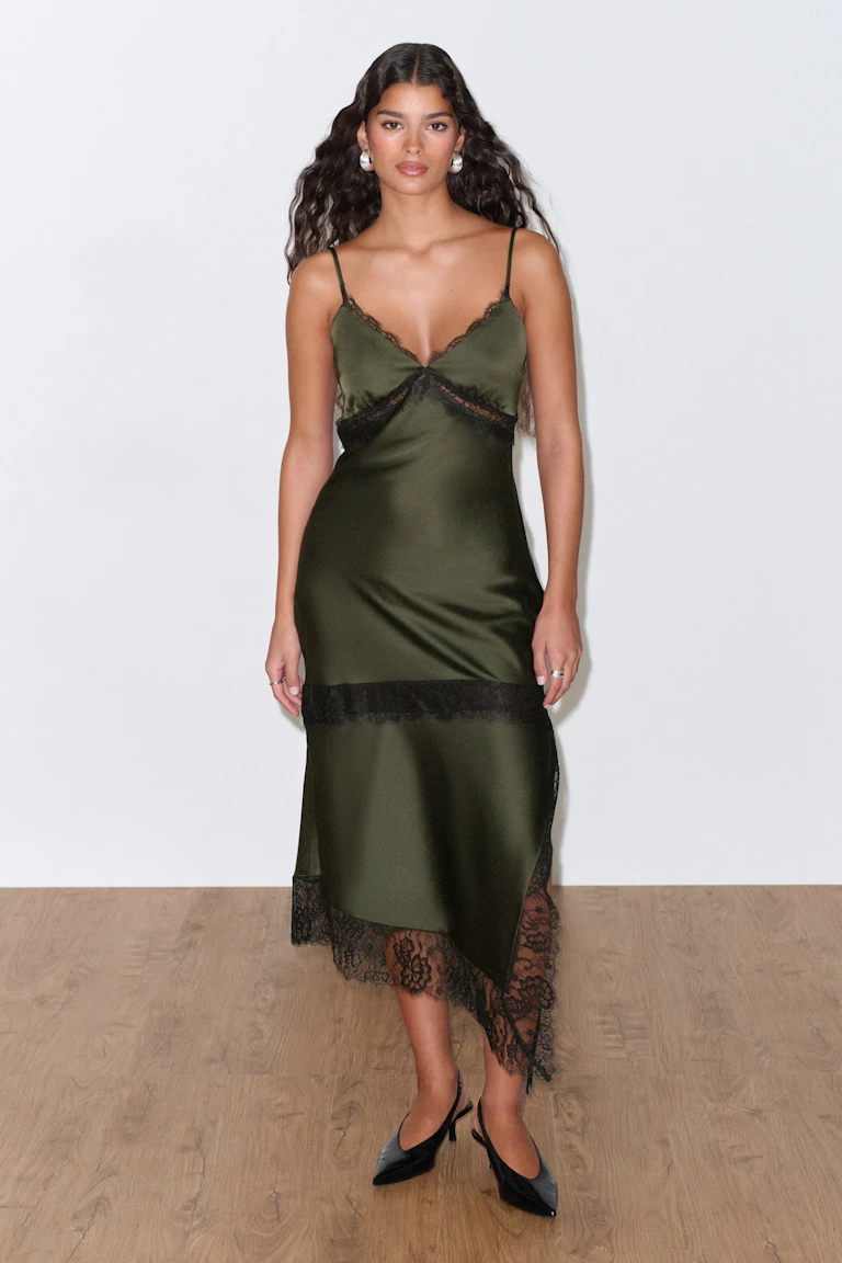 Olijfgroene satijnen slipjurk met dunne spaghettibandjes, V-halslijn en zwart kanten randje bij de buste, het middengedeelte en de asymmetrische zoom met kanten rand, gestyled met zwarte slingback-hakken met spitse teen en zilveren oorbellen.