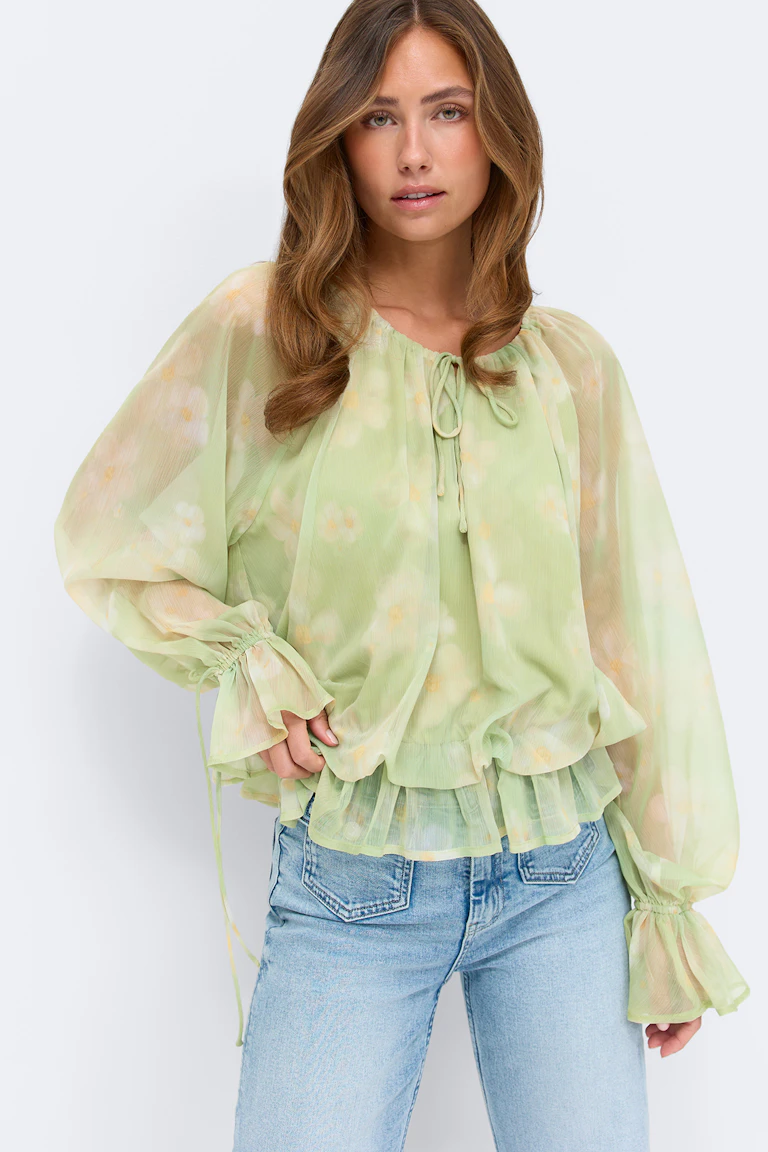 Blouse à manches longues transparente à imprimé floral vert pâle, avec encolure à nouer, ourlet à volants froncés et poignets à volants élastiques, associée à un jean clair.
