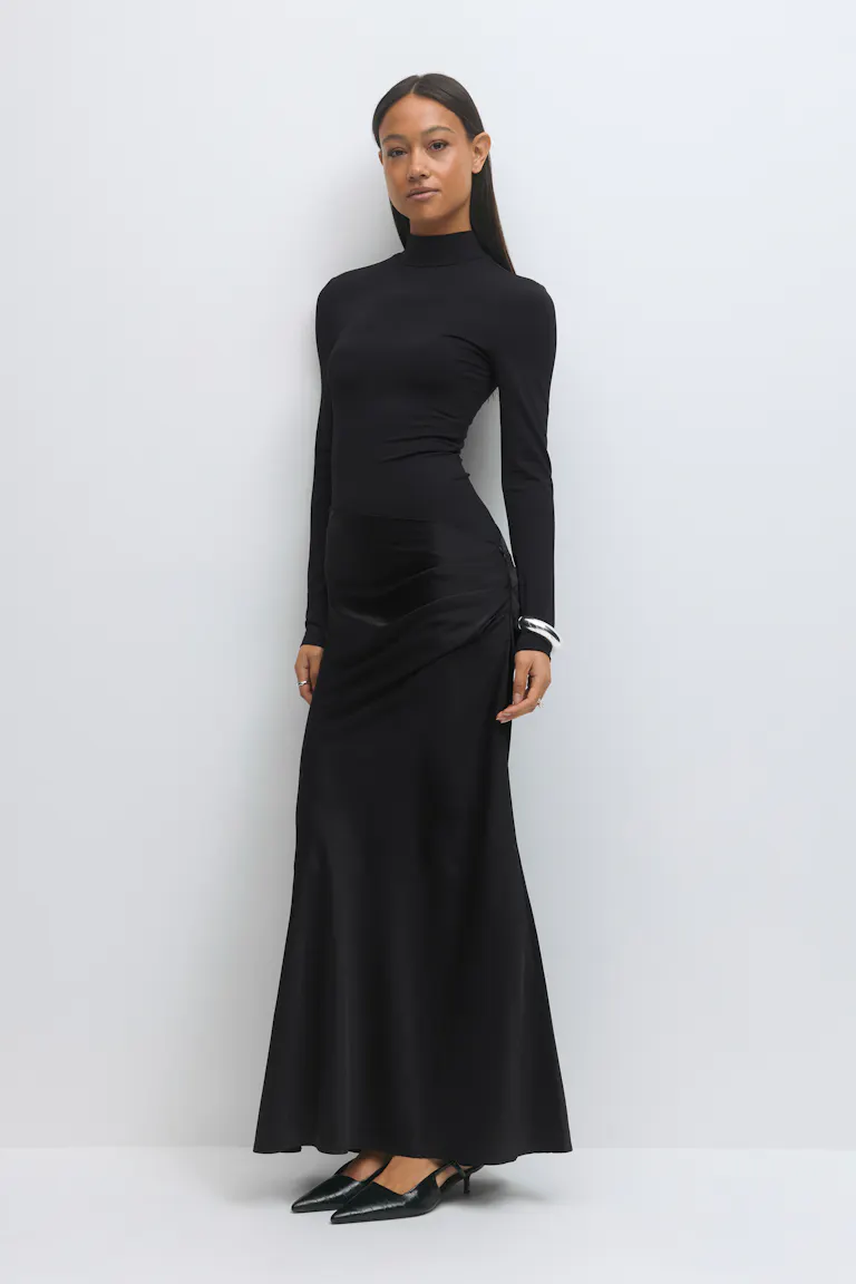 Schwarzes, tailliertes, langärmeliges Oberteil mit Rundhalsausschnitt, kombiniert mit einem schwarzen, gerüschten Maxirock aus Satin und schwarzen, spitz zulaufenden Slingback-Schuhen mit Kitten-Heel; silbernes Armband mit Manschette.