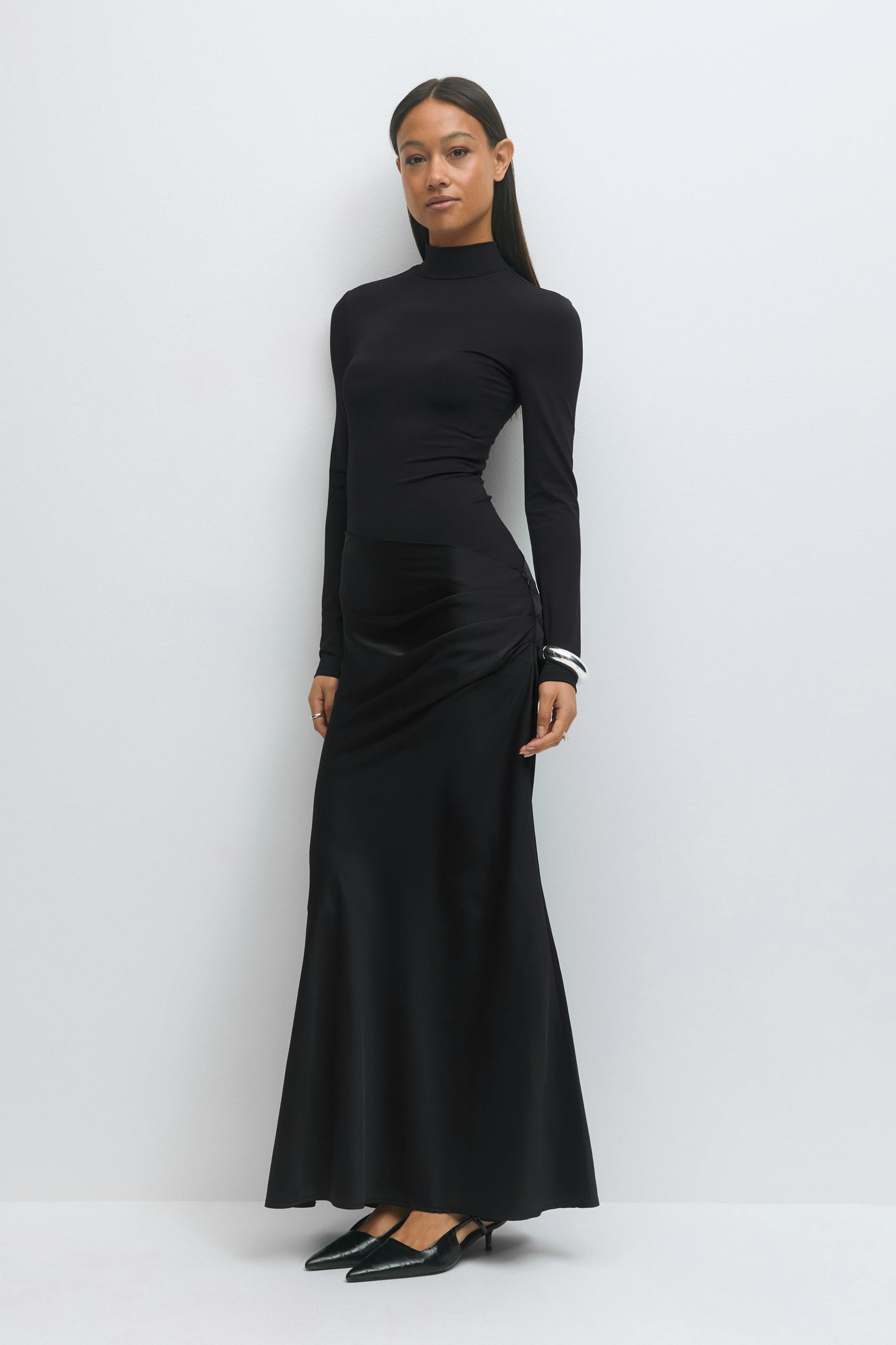 Fitted black long-sleeve mock-neck top paired with a black satin ruched maxi skirt and black pointed-toe kitten-heel slingback shoes; silver cuff bracelet.