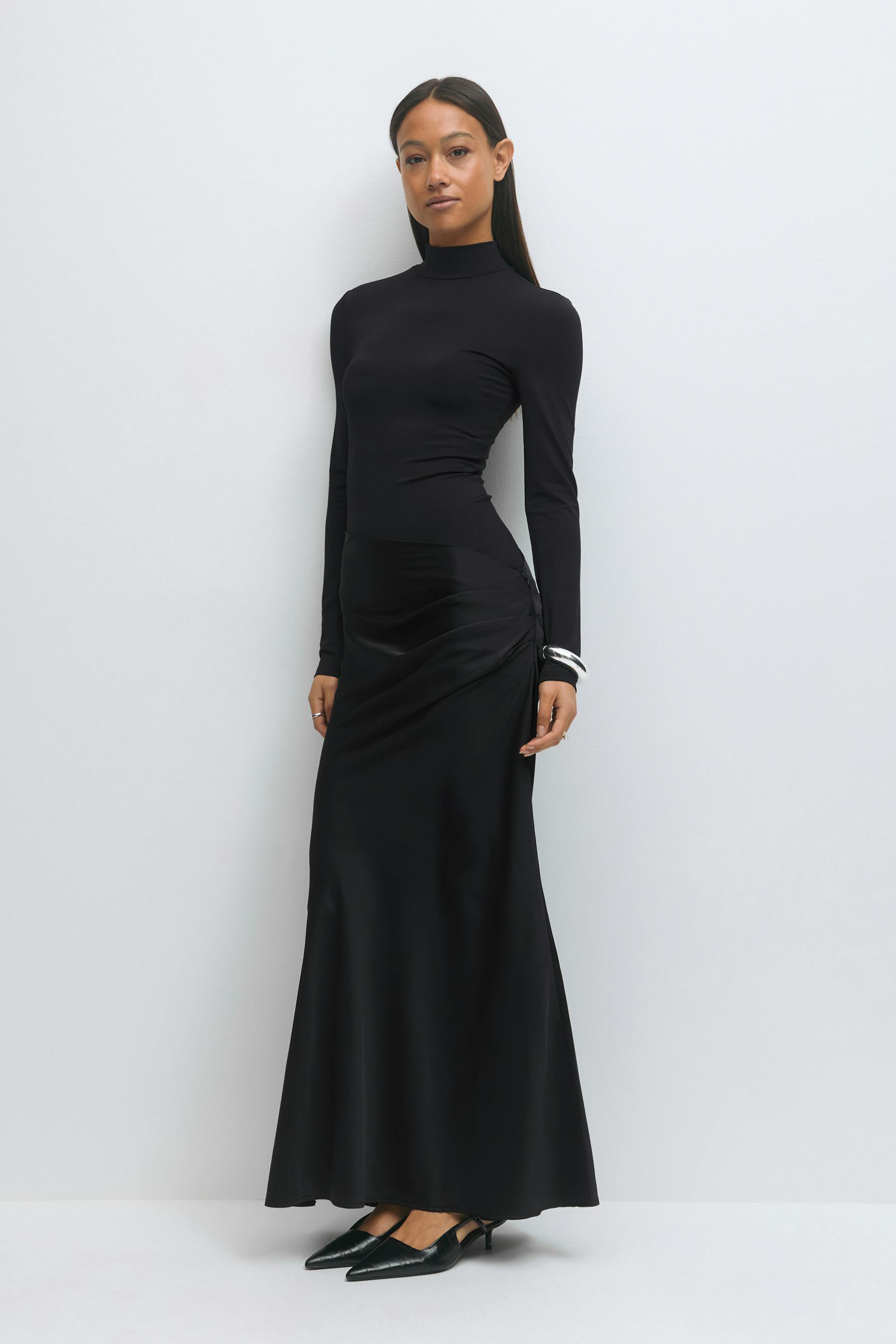 Fitted black long-sleeve mock-neck top paired with a black satin ruched maxi skirt and black pointed-toe kitten-heel slingback shoes; silver cuff bracelet.