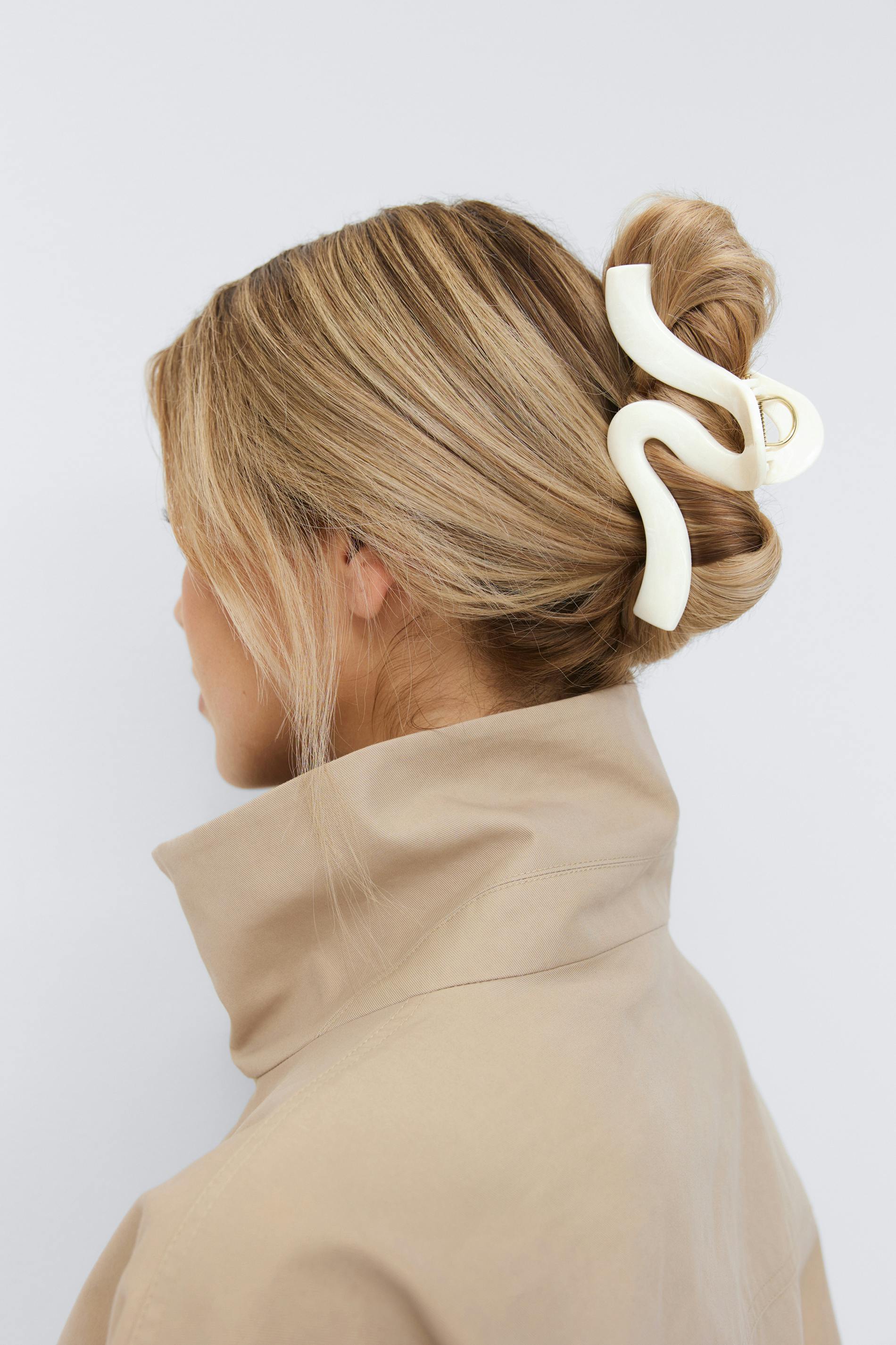 Beige frakke med høj krave kombineret med et elfenbensfarvet skulpturelt bølget hårspænde, der sikrer en lav knold.