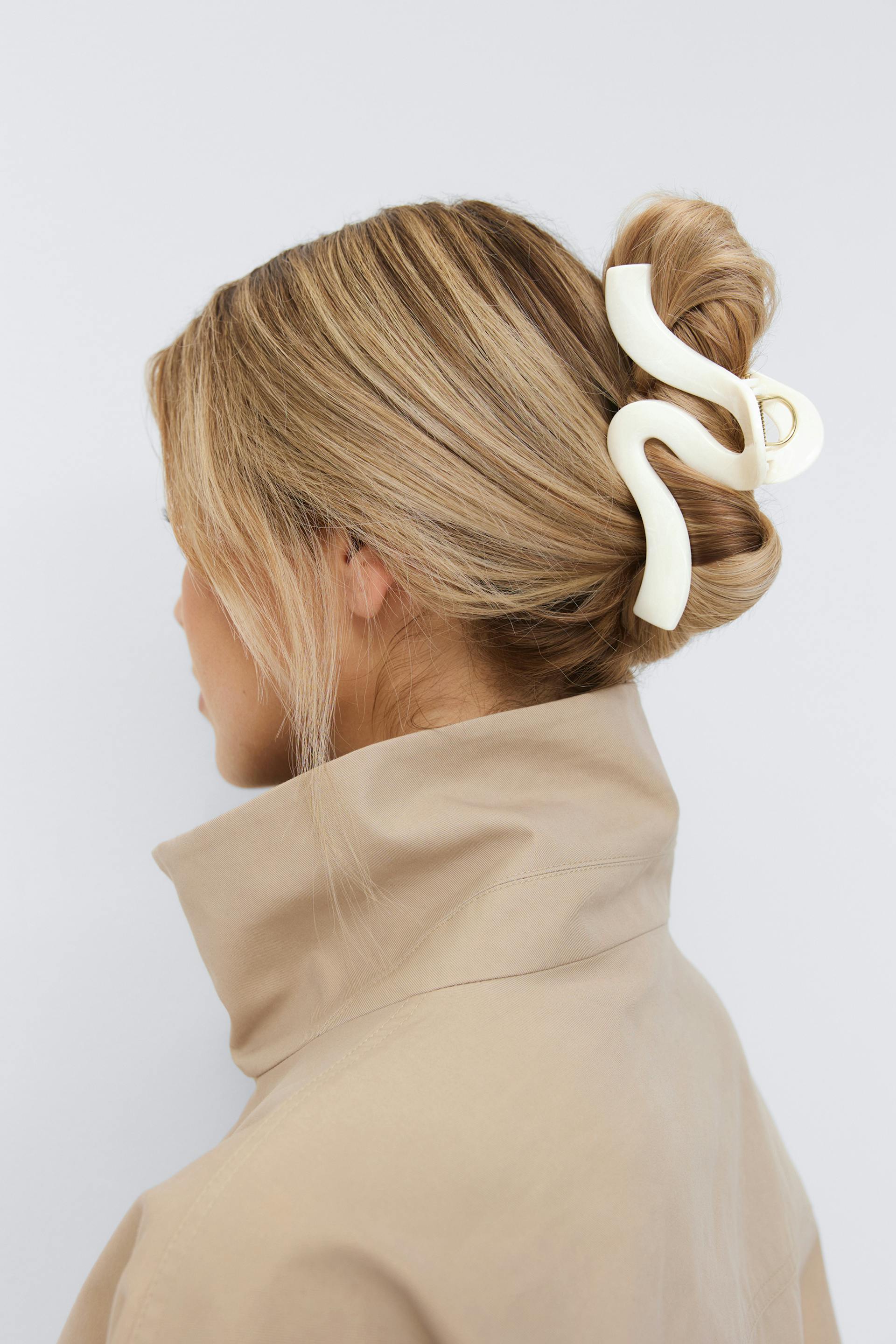 Beige frakke med høj krave kombineret med et elfenbensfarvet skulpturelt bølget hårspænde, der sikrer en lav knold.