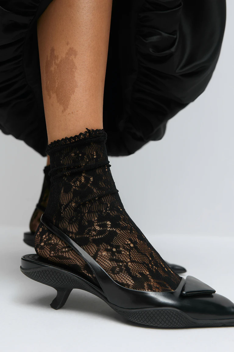 Svarta ankelstrumpor med blommig snörning tillsammans med svarta slingback-pumps i polerat läder, spetsiga tår och rektangulär vampdetalj.