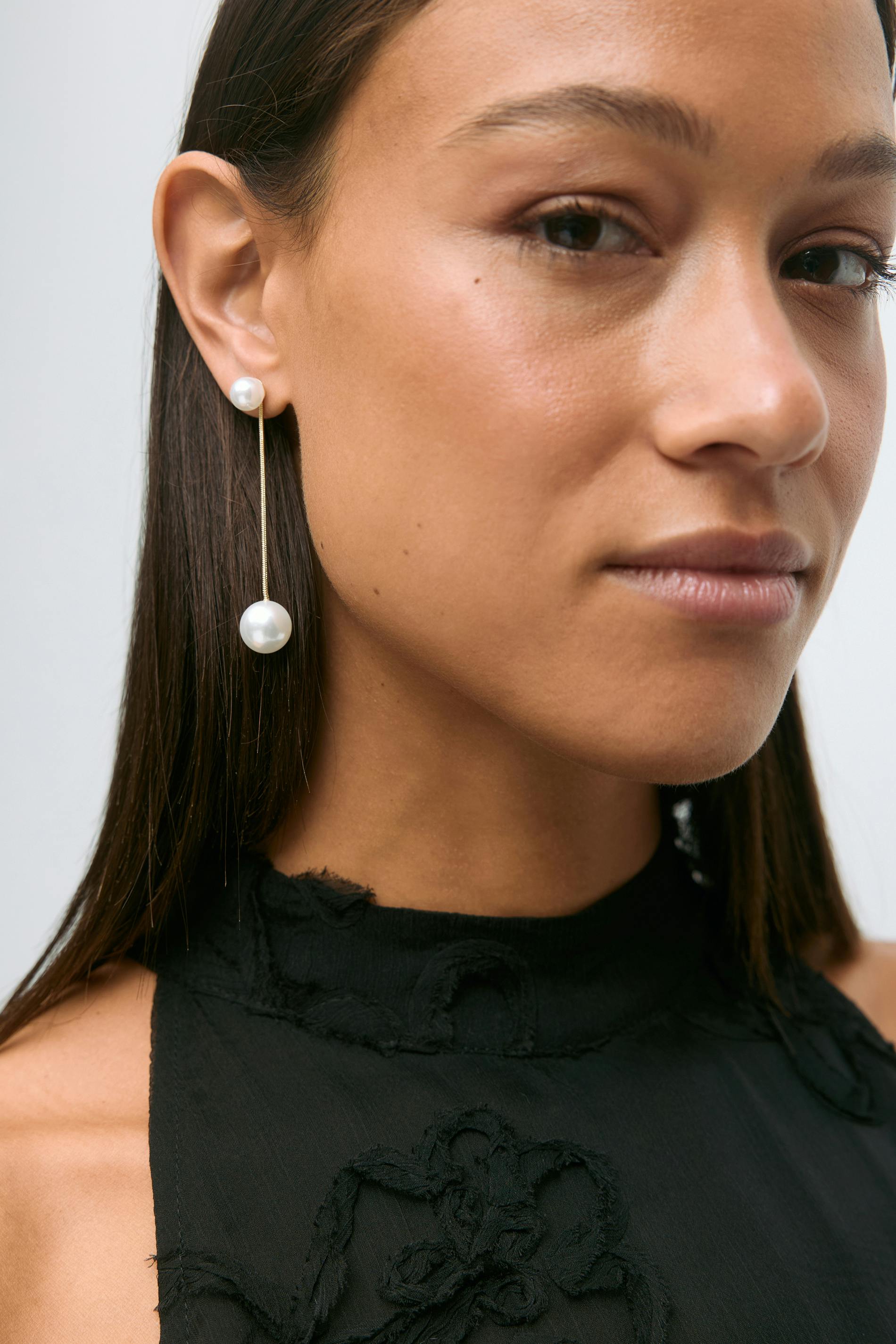 Schwarzes, hochgeschlossenes, ärmelloses Oberteil mit strukturierter Blumenapplikation und ausgefransten Rändern; lange Goldketten-Ohrringe mit einem kleinen Perlenstecker und einem größeren Perlenanhänger.