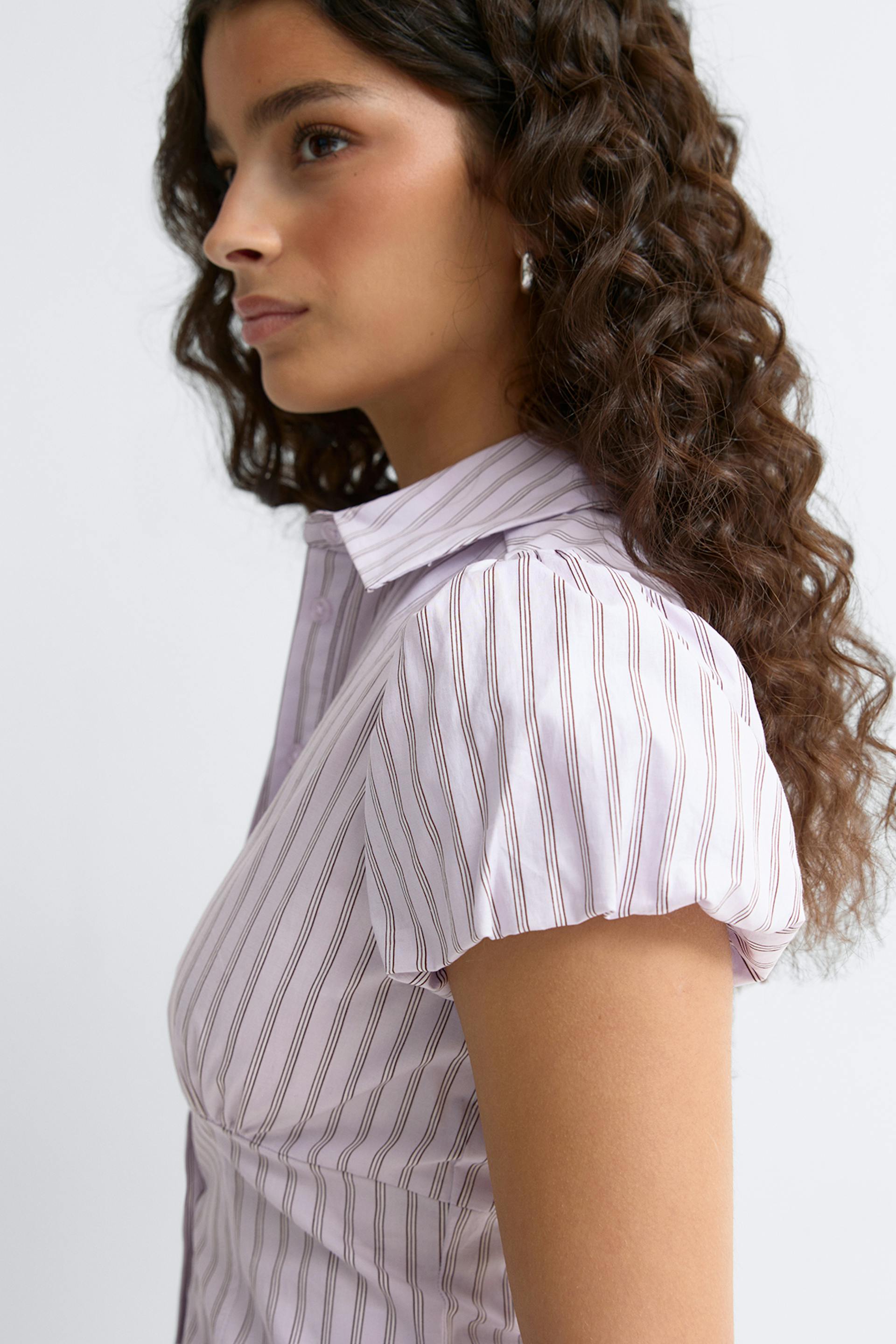 Vaalean laventelinvärinen napitettava pusero, jossa on ohuet pystyraidat, teräväkärkinen kaulus ja lyhyet pehmohihat; pieni hopeinen korvakoru.