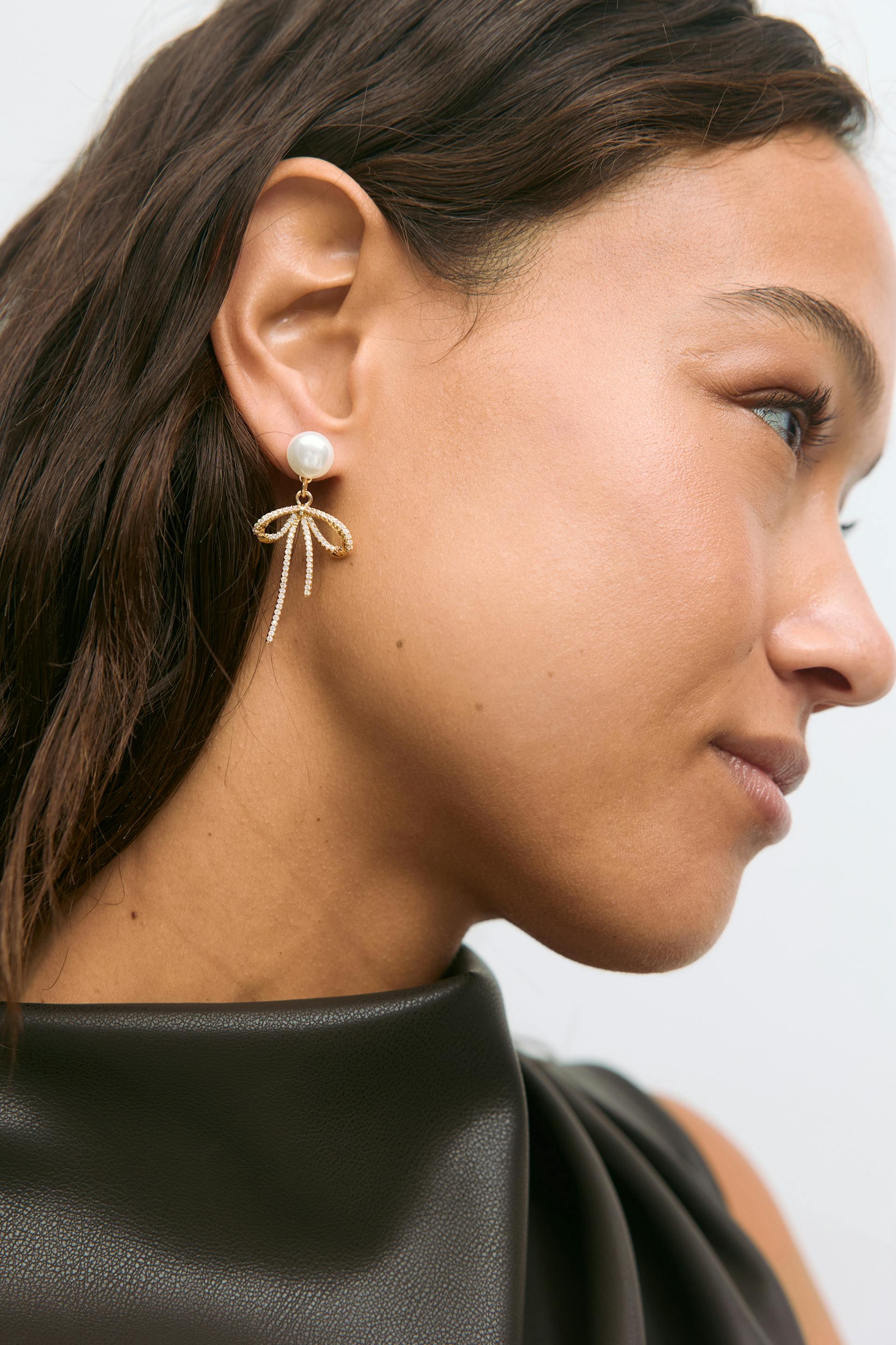 Black high-neck leather-look top and gold-tone drop earrings featuring a pearl stud and bow-shaped, rhinestone-encrusted dangling strands.