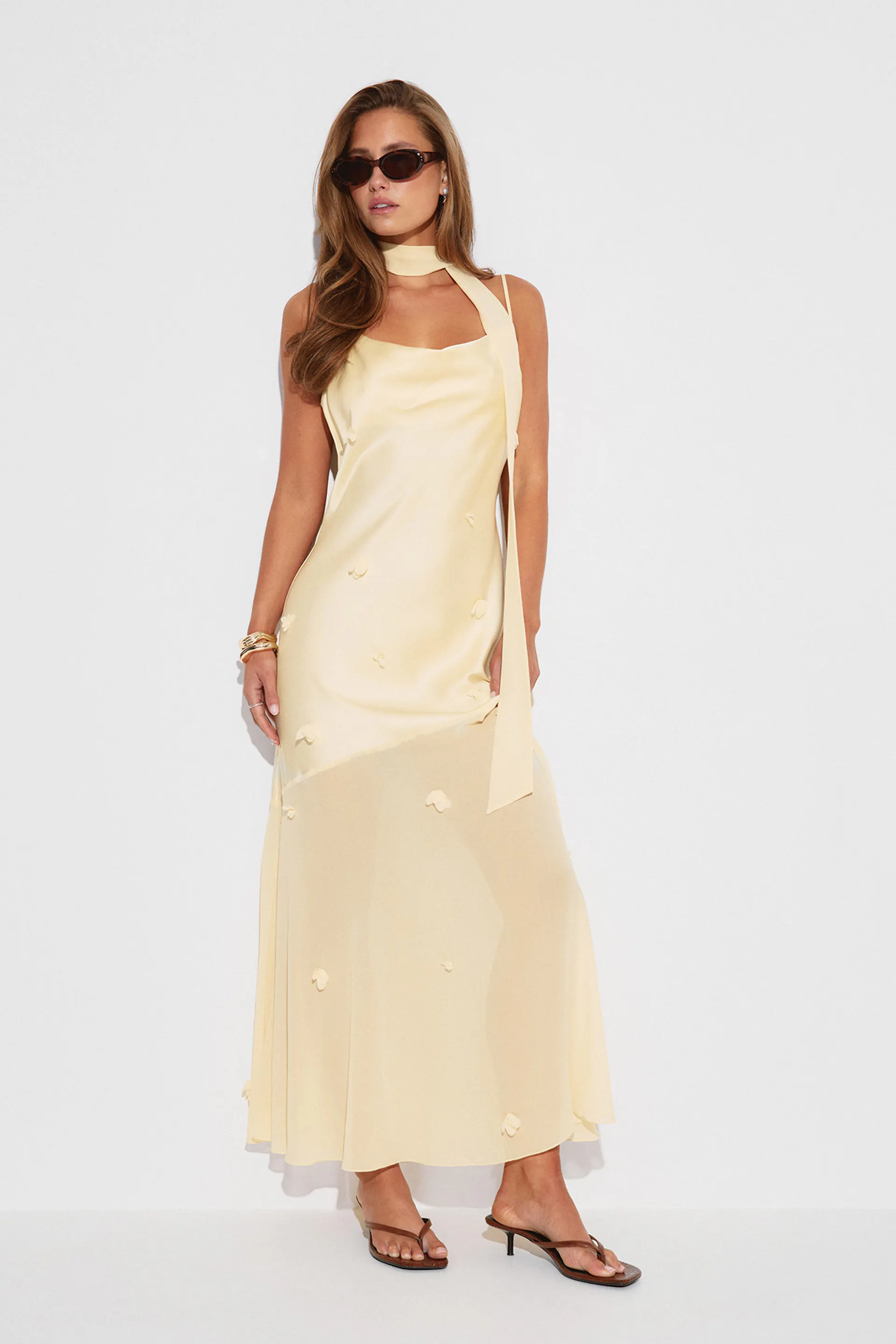 Robe longue en satin jaune pâle à bretelles spaghetti avec un long foulard attaché au cou, des applications florales éparses et une jupe inférieure légèrement transparente, associée à des sandales à talons brunes, des lunettes de soleil ovales foncées et des bracelets en or.