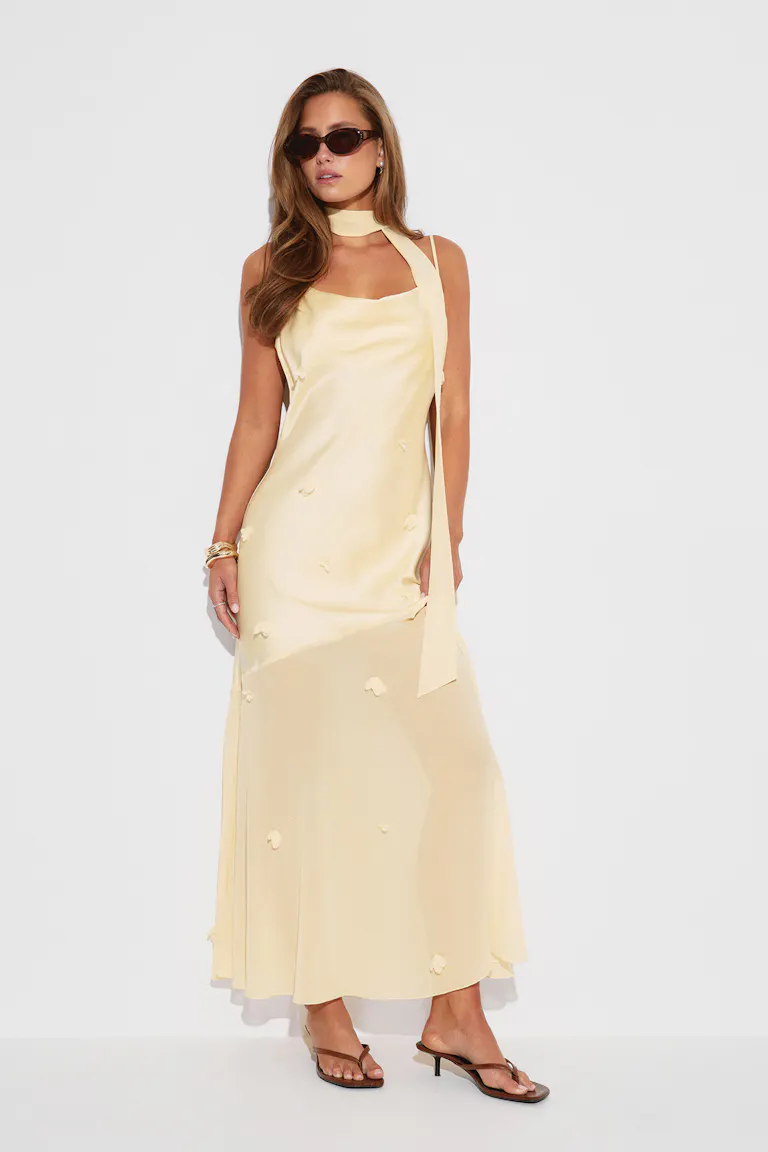 Hellgelbes Maxischlupfkleid aus Satin mit Spaghettiträgern und langem Halstuch, verstreuten Blumenapplikationen und einem leicht durchsichtigen unteren Rockteil, kombiniert mit braunen Zehenstegsandalen, einer dunklen ovalen Sonnenbrille und goldenen Armbändern.