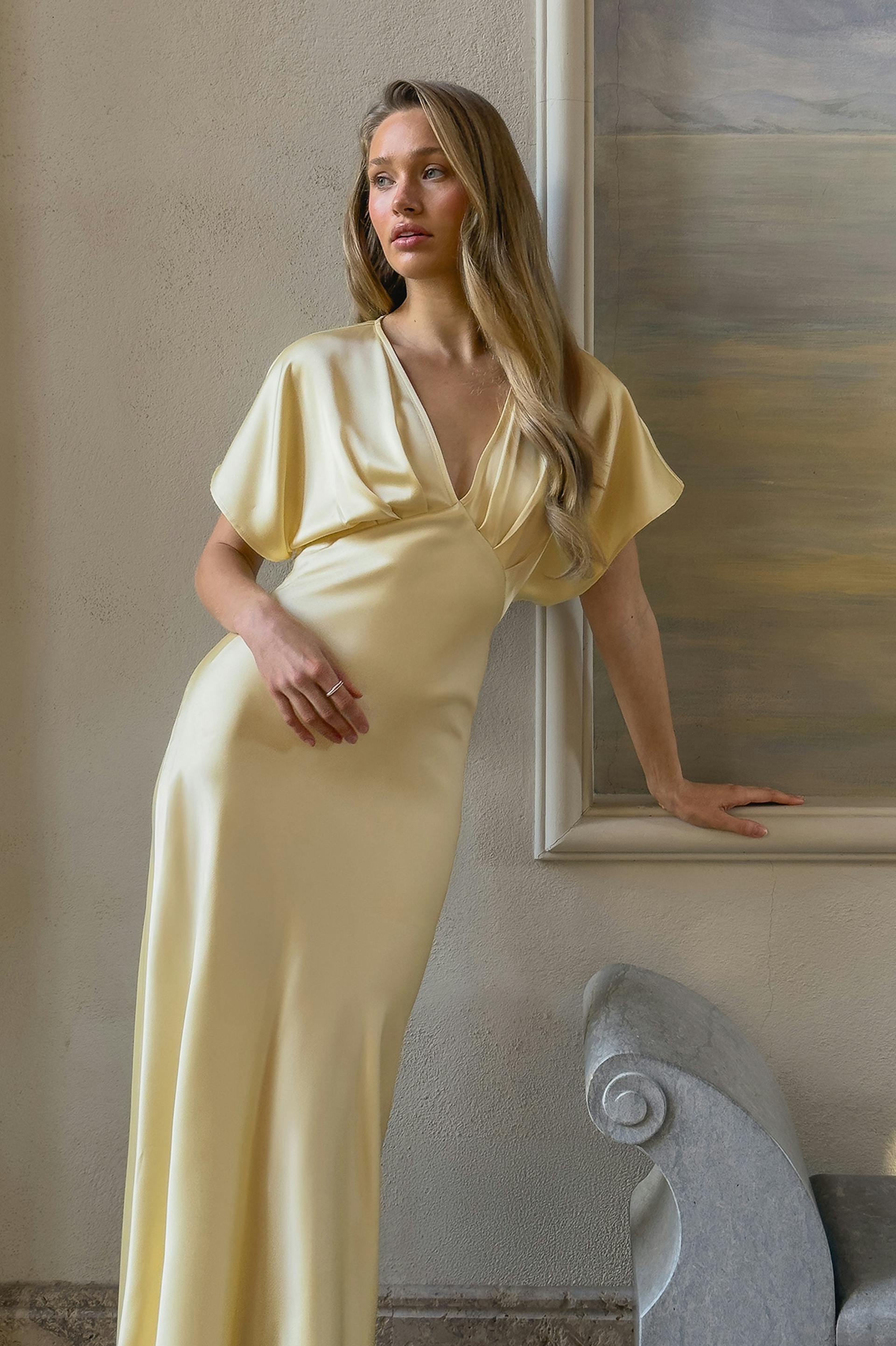 Lattianmyötäinen vaaleankeltainen satiinipuku, jossa on syvä V-pääntie, drapattu laskostettu liivi, lyhyet kimonohihat ja istuva vyötärö.