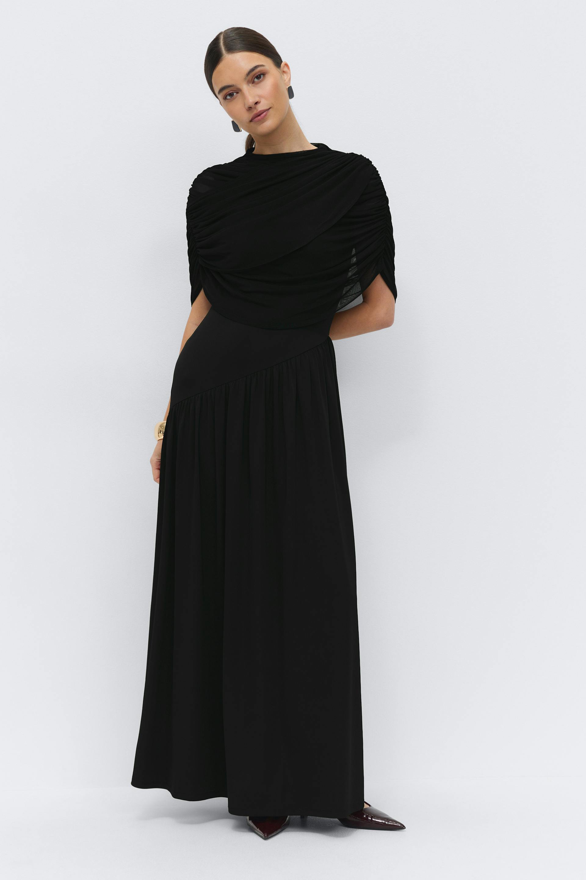 Black floor-length gown with a ruched, draped overlay and fitted waist; styled with black stud earrings, a gold cuff bracelet and pointed burgundy heels.