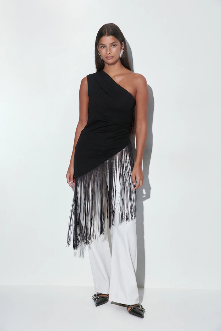 Schwarzes, asymmetrisches One-Shoulder-Oberteil mit langen schwarzen Fransen, weiße Jeans mit weitem Bein, schwarze, spitz zulaufende Riemenflats mit silbernen Schnallen und silberne Statement-Ohrringe.