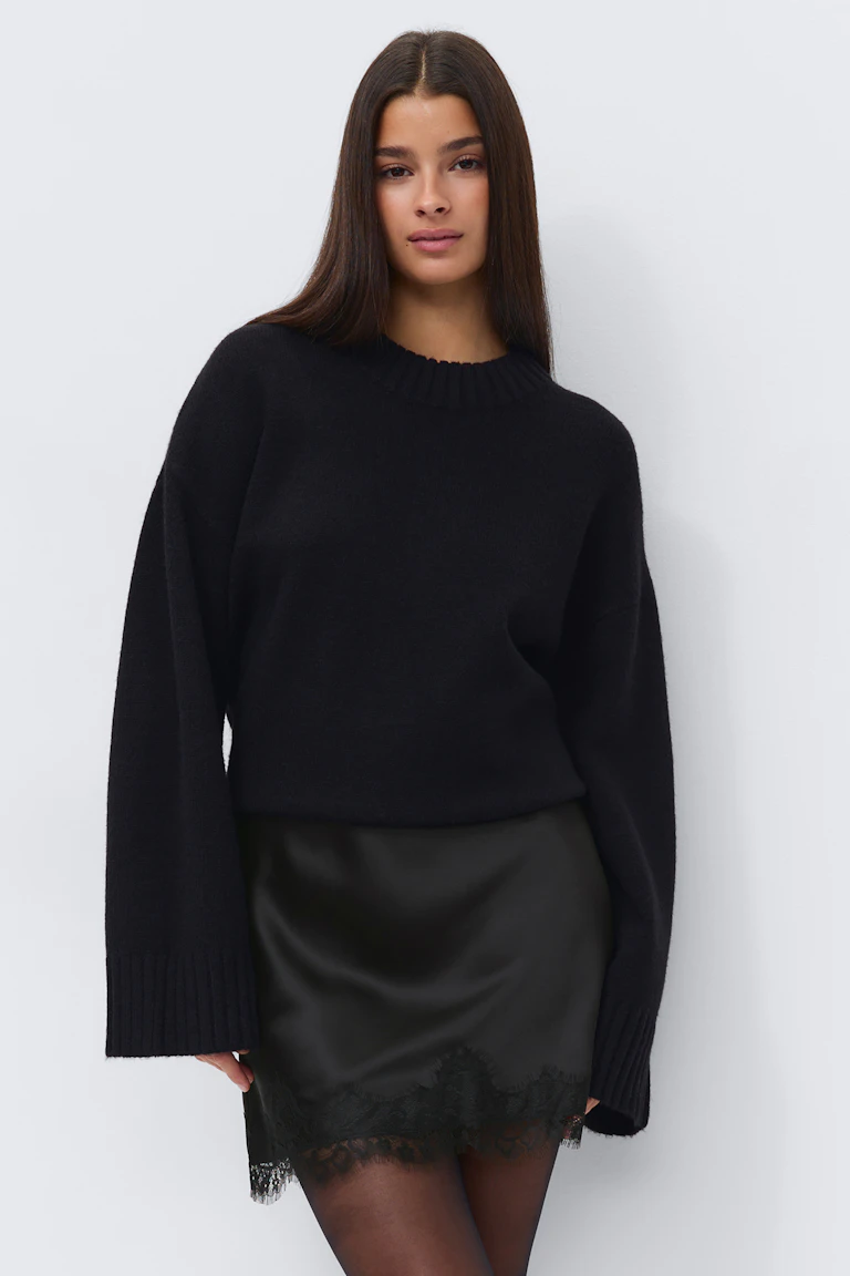 Schwarzer Oversize-Pullover aus Rippstrick mit verlängerten Bündchen, schwarzer Satin-Minirock mit Spitzenbesatz am Saum und schwarze Strumpfhose.