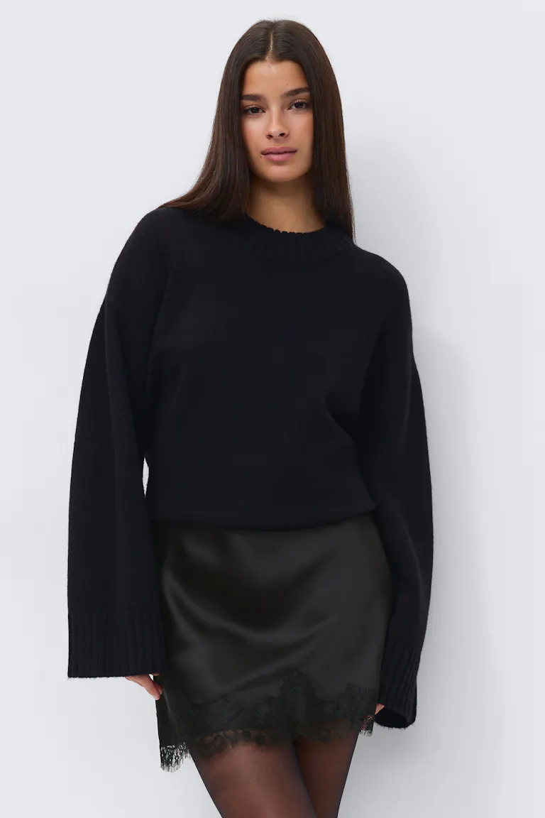 Sort oversized sweater i ribstrik med forlængede manchetter, sort mininederdel i satin med blondekant forneden og gennemsigtige sorte strømpebukser.