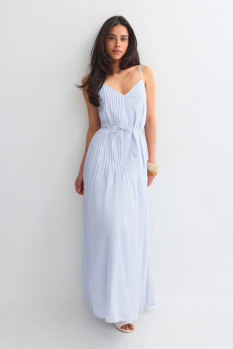 Light blue pleated maxi dress with thin spaghetti straps, V-neckline and a self-tie waist, paired with white heeled thong sandals.