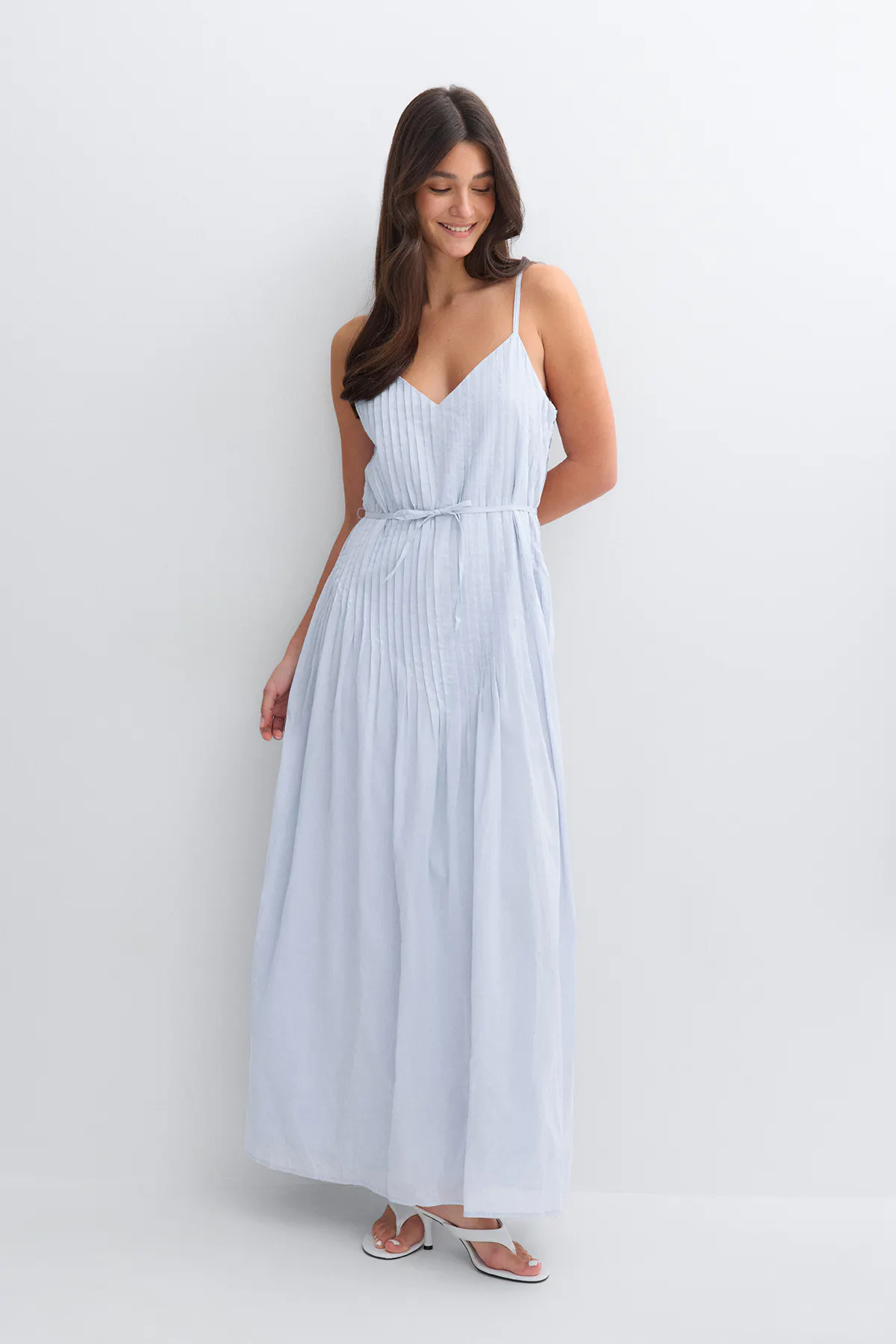 Light blue pleated maxi dress with thin spaghetti straps, V-neckline and a self-tie waist, paired with white heeled thong sandals.