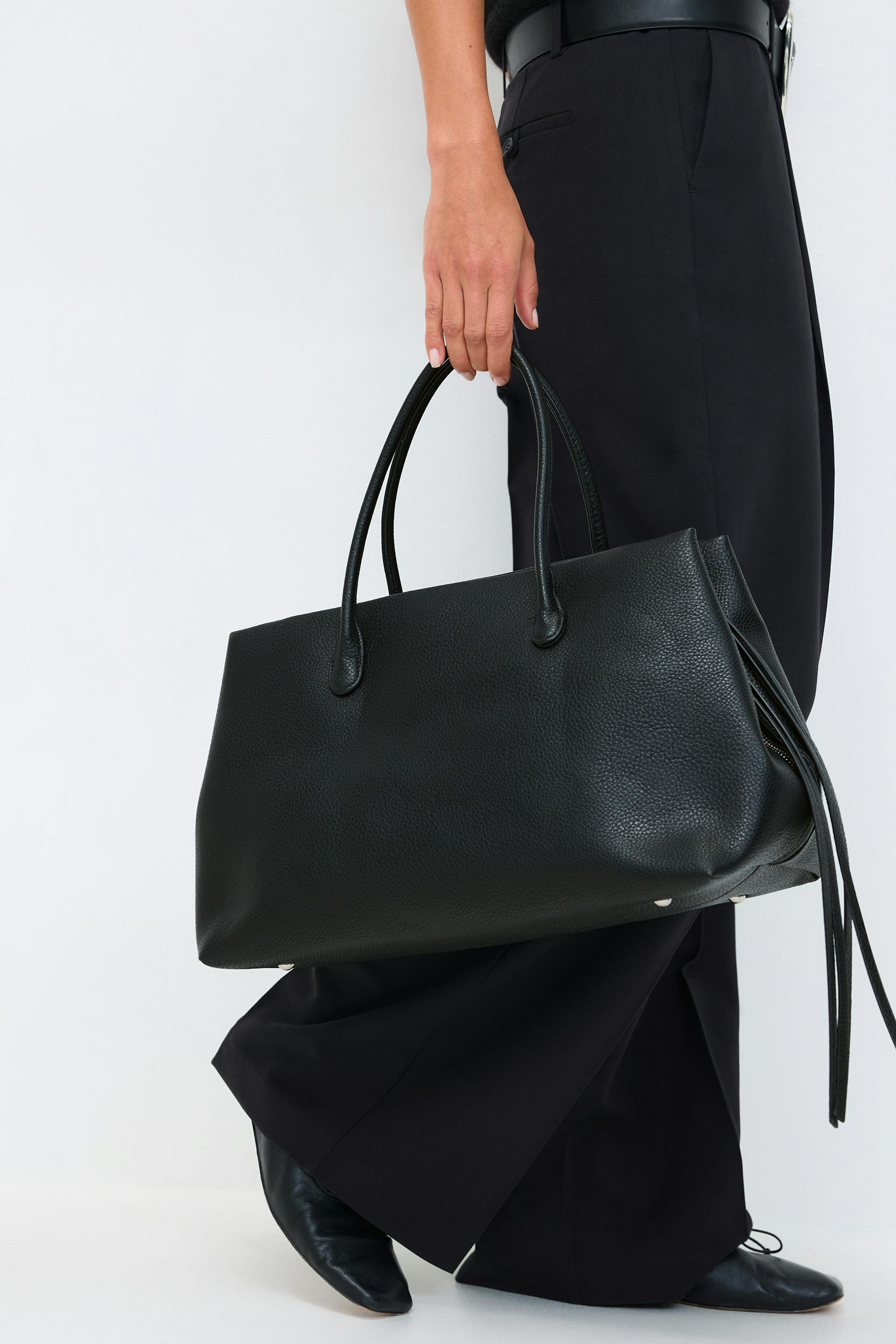Black wide-leg trousers with a black leather belt, paired with black pointed-toe leather flats and a large black pebbled-leather tote bag with double top handles and tassel detail.