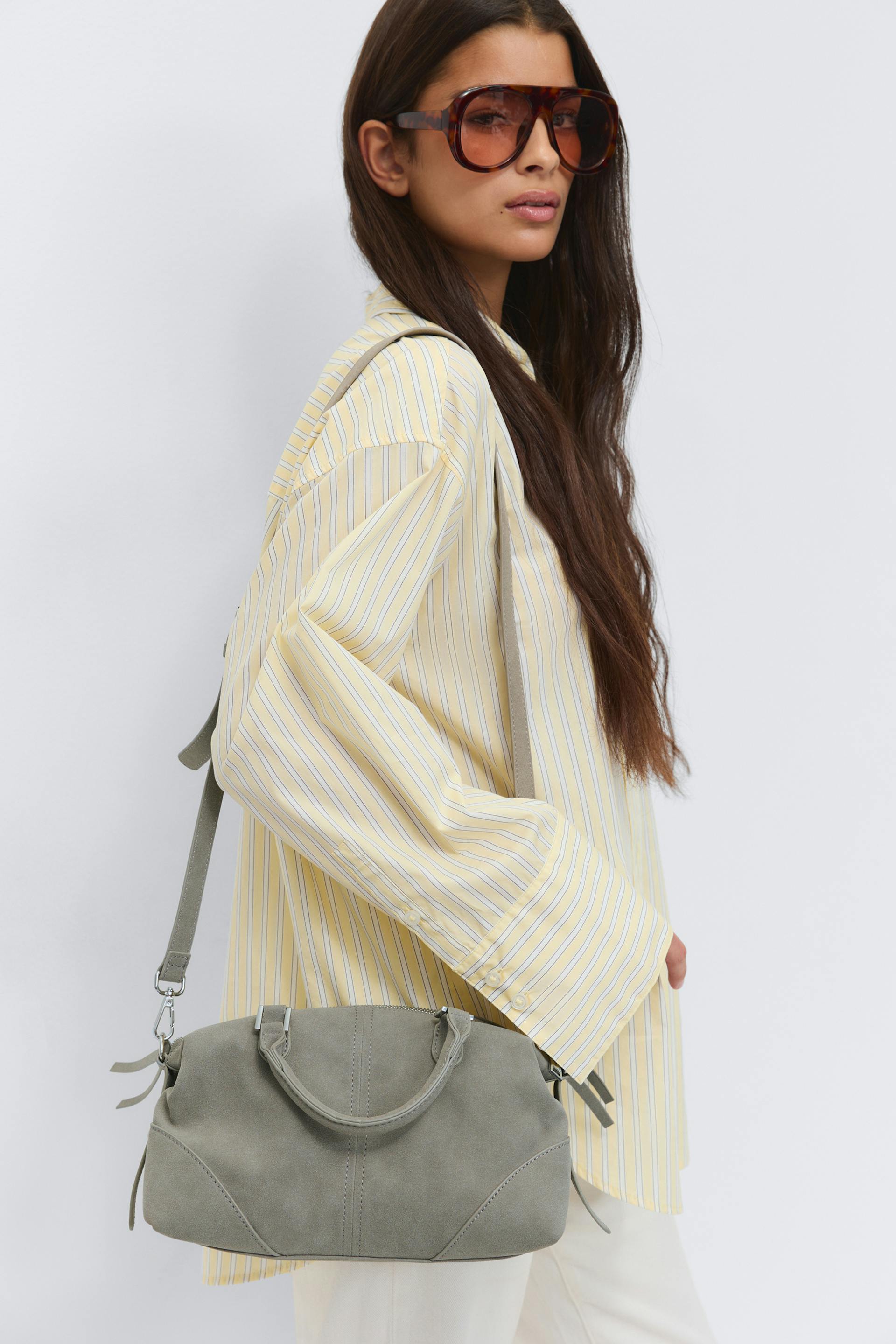 Oversized yellow-and-white striped button-down shirt, cream trousers, tortoiseshell oversized sunglasses, and a grey suede handbag with top handles and an adjustable shoulder strap.