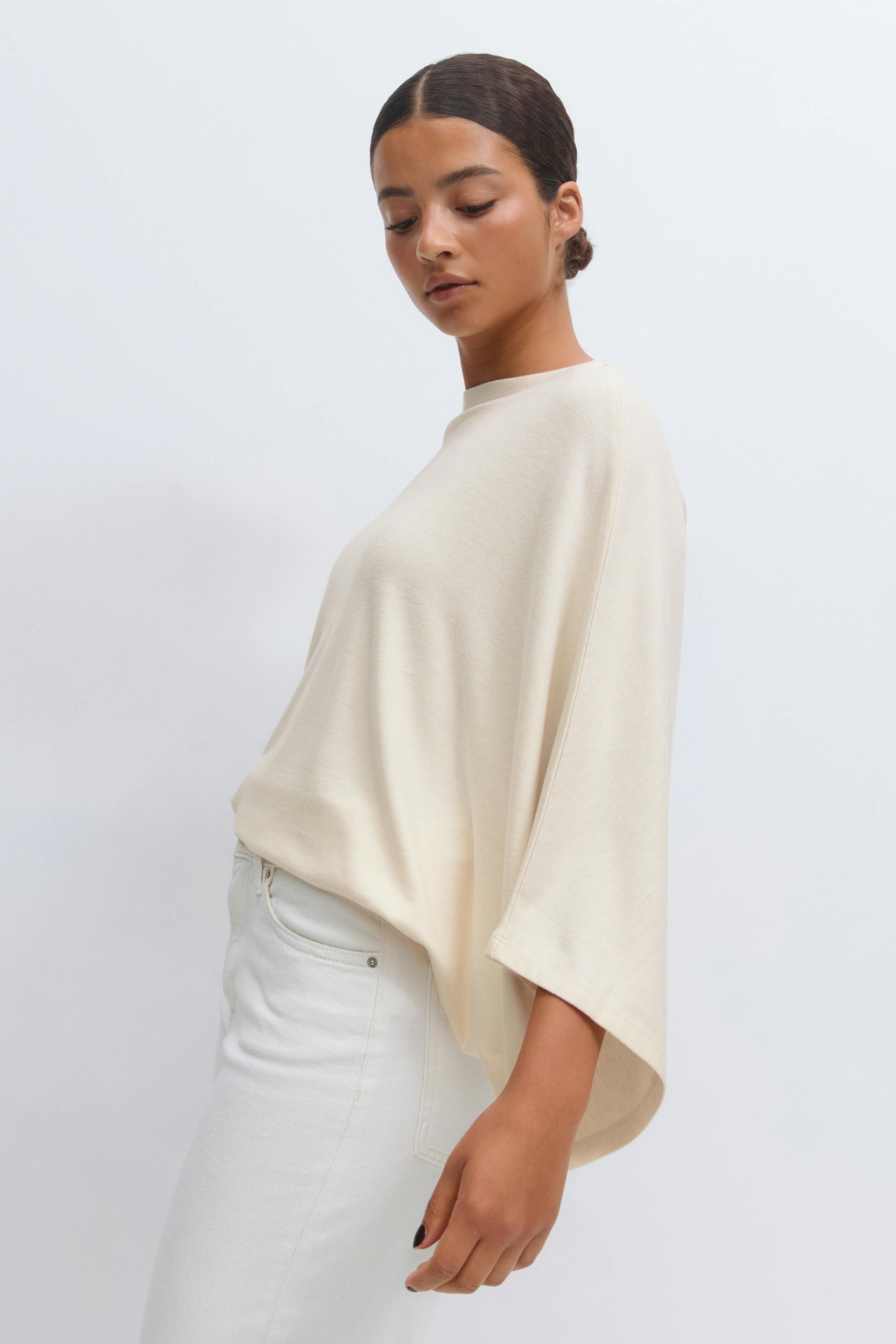 Cream oversized top with wide elbow-length sleeves and a high round neckline, paired with high-waisted white jeans.