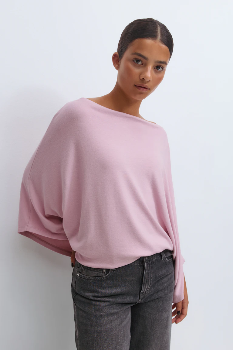 Lys rosa, oversized strikketopp med båthals og dolmanermer, delvis stukket ned i høytaljede, falmede jeans i svart/kull.