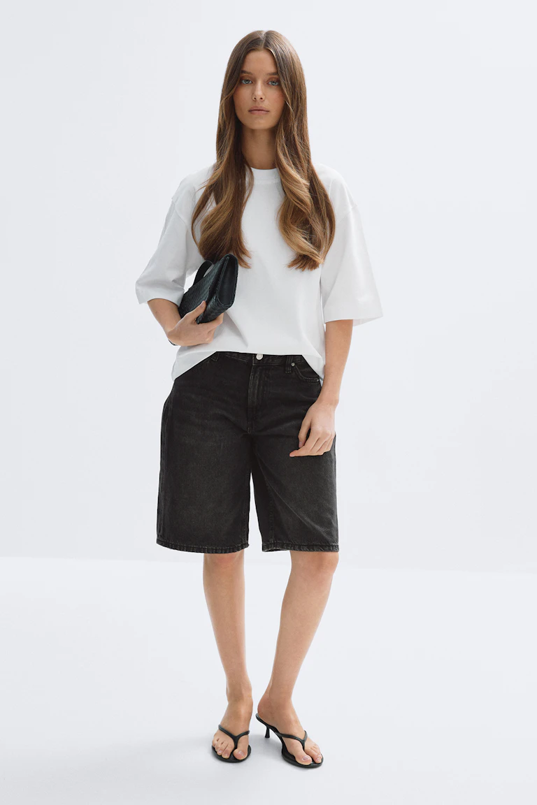 Wit T-shirt met oversized korte mouwen in combinatie met een zwarte denim short met knielengte, zwarte teenslippers met hak en een zwarte clutch.