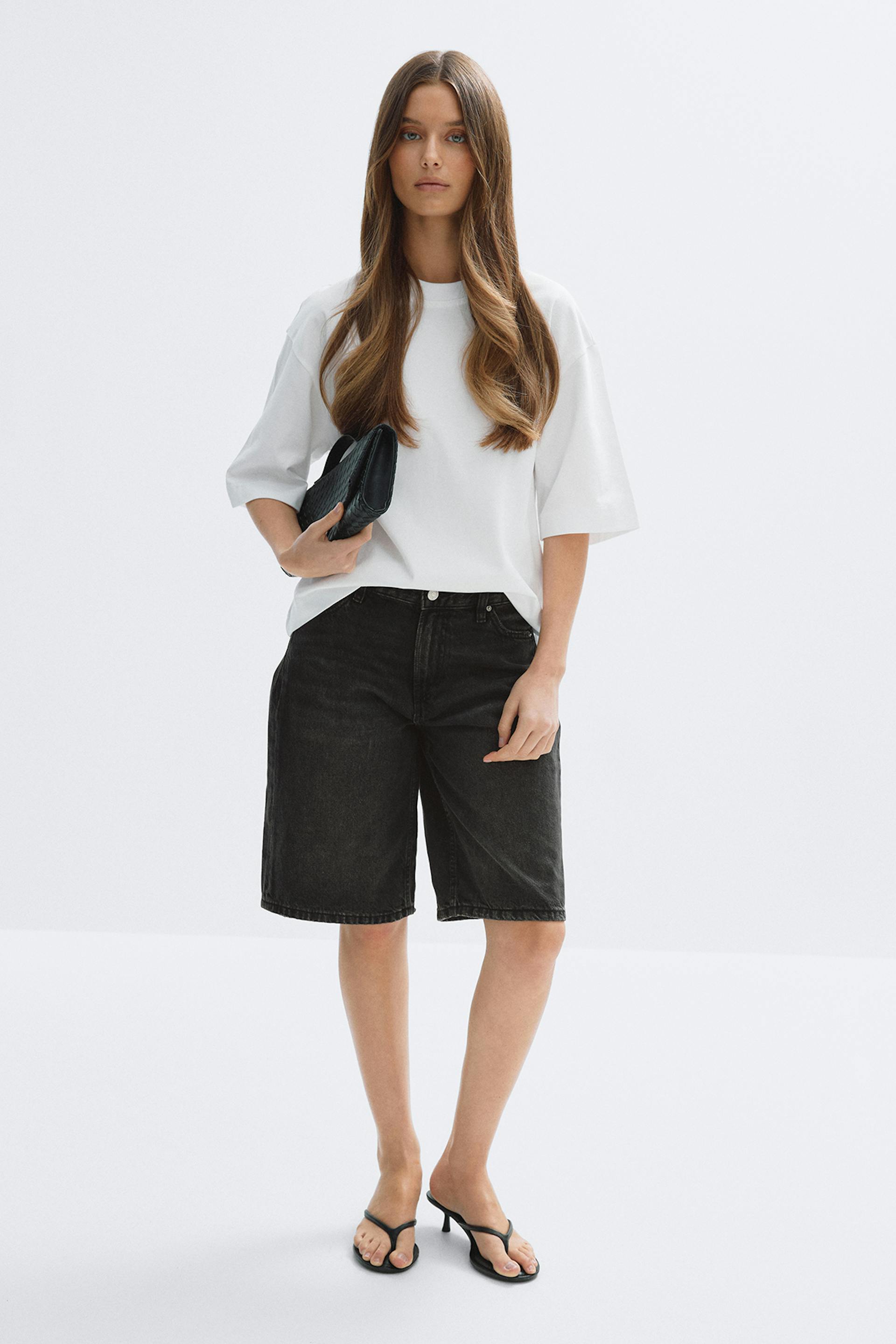 T-shirt blanc surdimensionné à manches courtes associé à un short en denim noir au genou, des sandales à talons noires et une pochette noire.