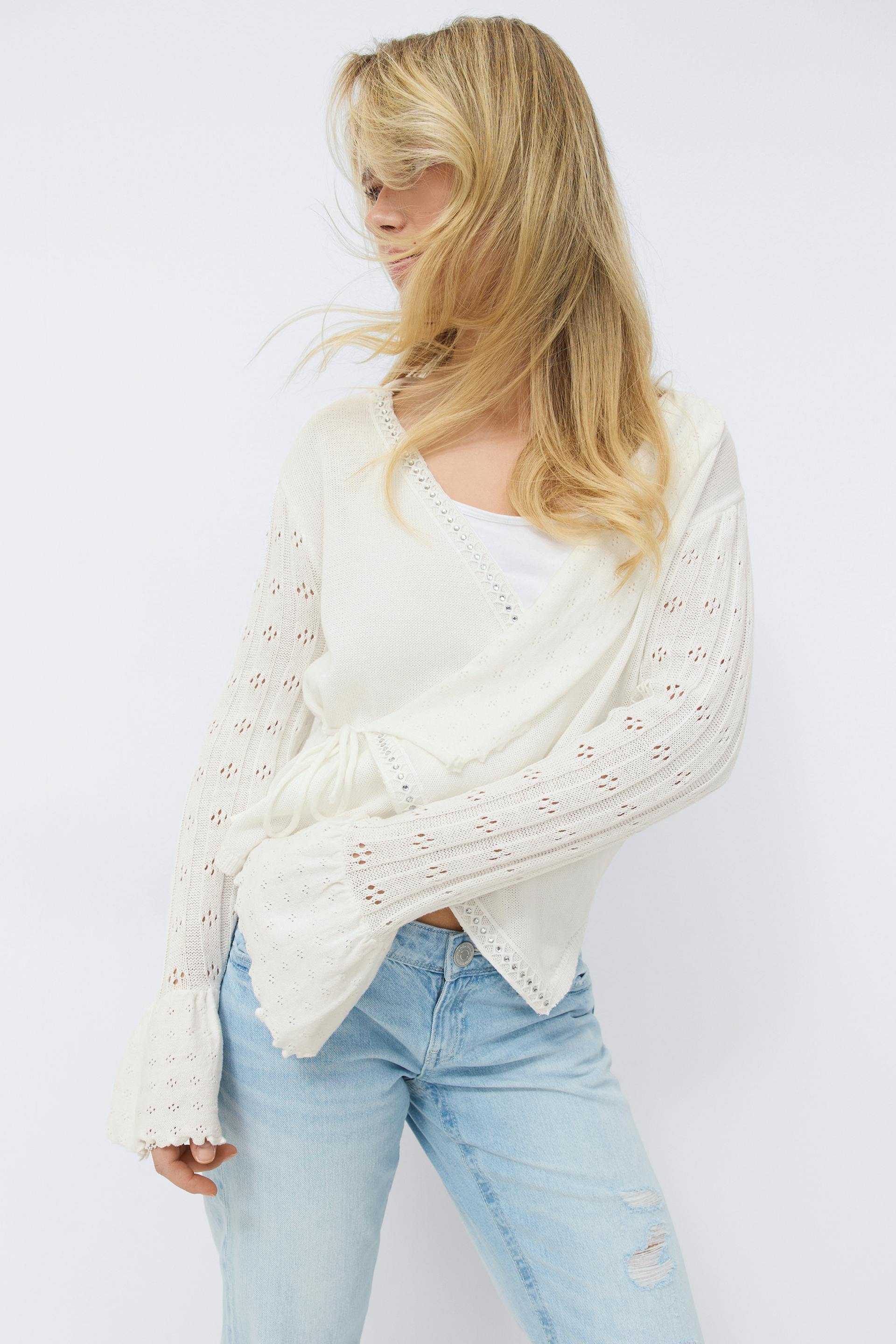 Cream wrap-style eyelet knit cardigan with tie waist and flared eyelet sleeves, worn over a white tank top and paired with light-wash distressed jeans.