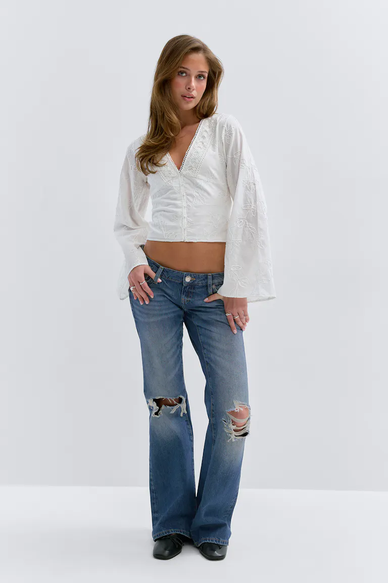 Witte geborduurde blouse met V-hals en kanten rand en lange klokmouwen; medium-wash distressed flared jeans met gescheurde knieën; zwarte enkellaarsjes met spitse teen.