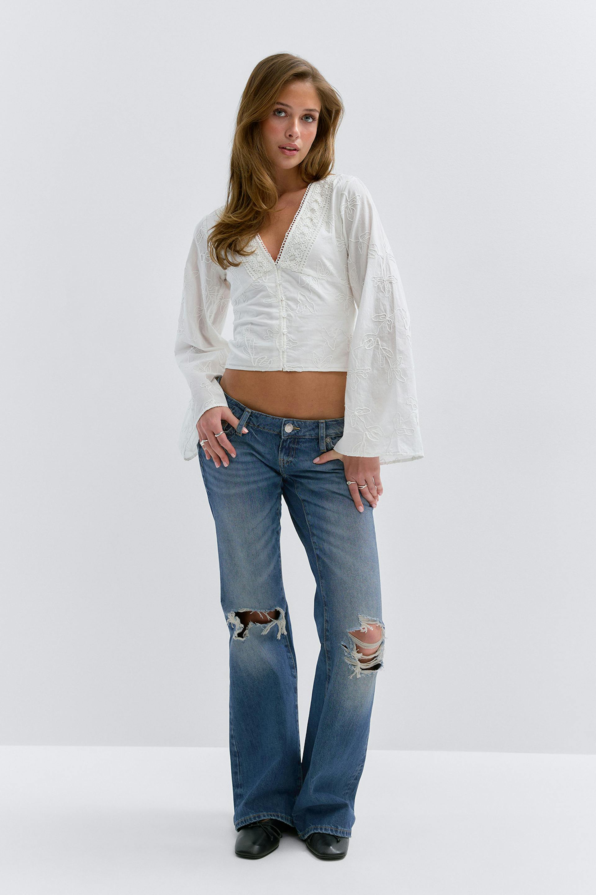 Weiße, bestickte Bluse mit V-Ausschnitt, Spitzenbesatz und langen Glockenärmeln; mittelschwere Jeans mit zerrissenen Knien; schwarze, spitz zulaufende Ankle Boots.