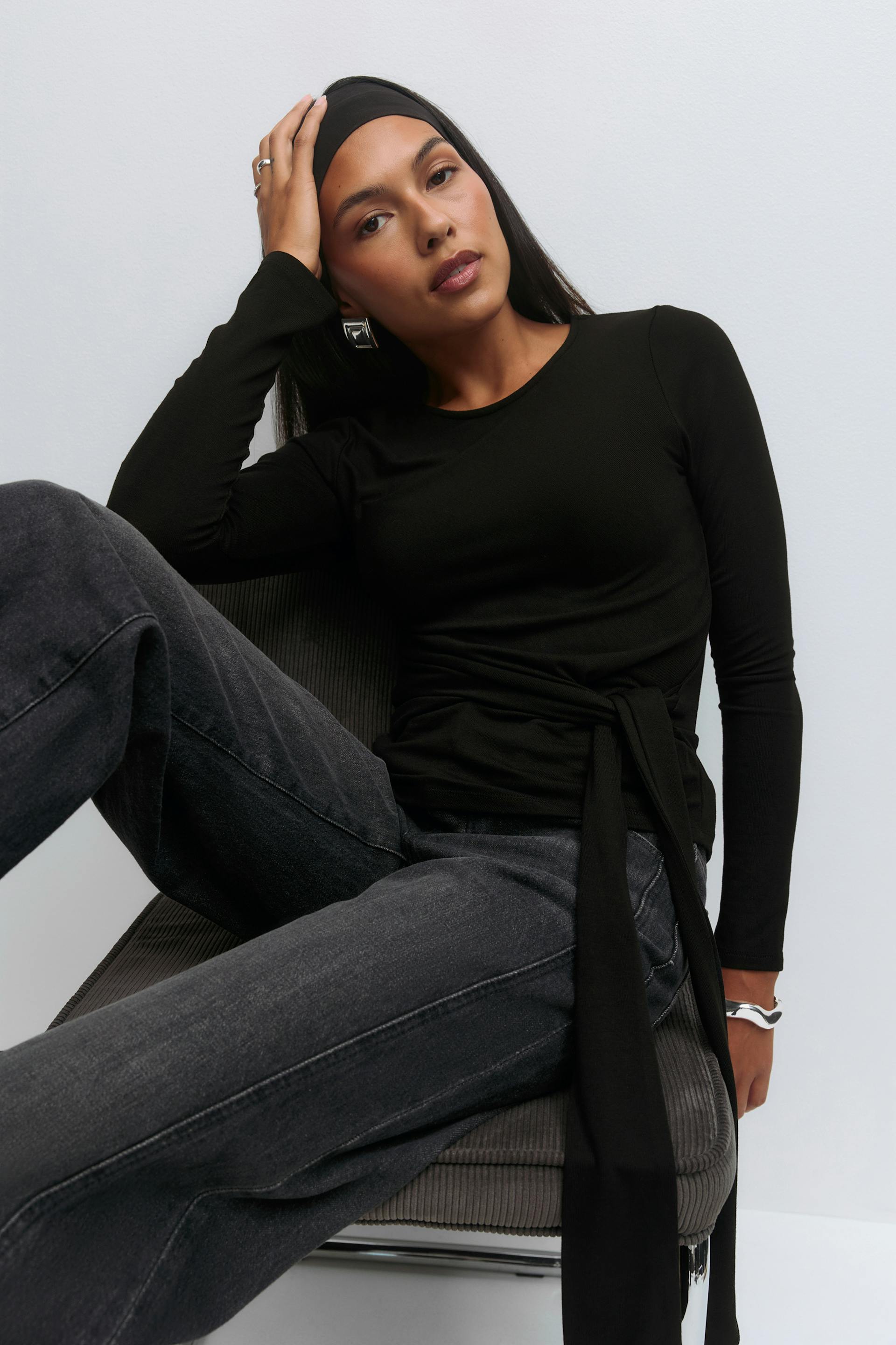 Schwarzes langärmeliges Oberteil mit Rundhalsausschnitt und Wickelgürtel, schwarzes Stirnband, verblasste schwarze Jeans mit weitem Bein, silberne geometrische Reif-Ohrringe, silbernes Armband und silberne Ringe.