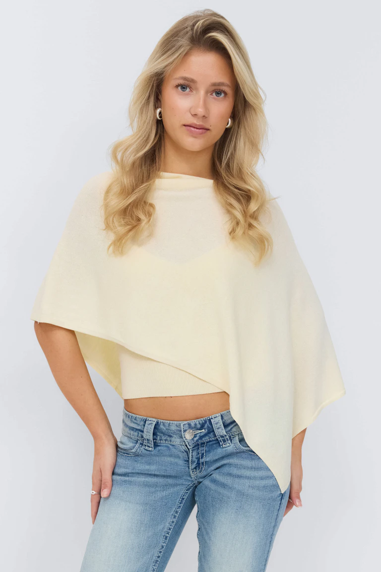 Lichtgele asymmetrische poncho in laagjes over een bijpassende geribde top, gecombineerd met een jeans met lichte taille.