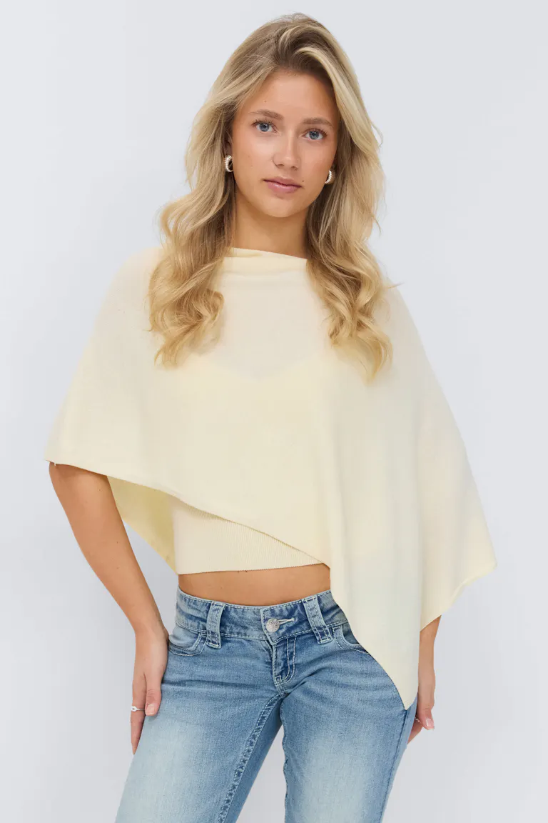 Lichtgele asymmetrische poncho in laagjes over een bijpassende geribde top, gecombineerd met een jeans met lichte taille.